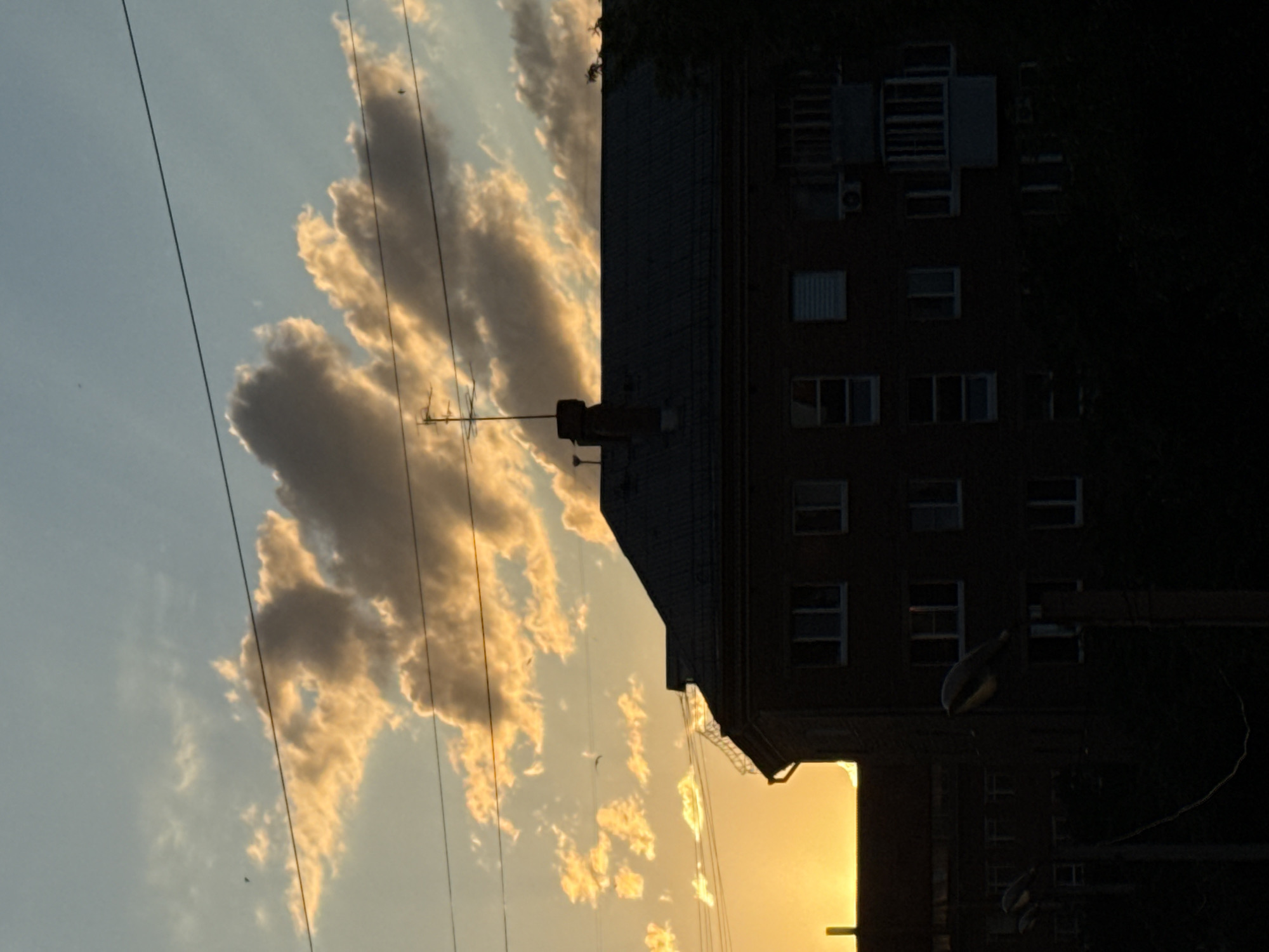The image is a photograph taken at sunset in Yekaterinburg, Russia. The scene is dominated by a building, silhouetted against a sky filled with dramatic clouds illuminated by the setting sun. The clouds are a mix of light yellow and grey, suggesting a dynamic weather pattern. Several power lines crisscross the sky, adding a sense of urban life to the natural beauty of the sunset.
The building appears to be an apartment block, with numerous windows visible, some reflecting the warm light of the sunset. Various air conditioning units are mounted on the exterior, indicating a residential setting. A tall antenna stands on the roof of the building, further suggesting an urban environment. The lower portion of the image is darker, with the warm glow of the setting sun visible in the lower right corner, partially obscured by the building. The overall mood is one of quiet observation, capturing the transition from day to night in a city. There is no visible text in the image.