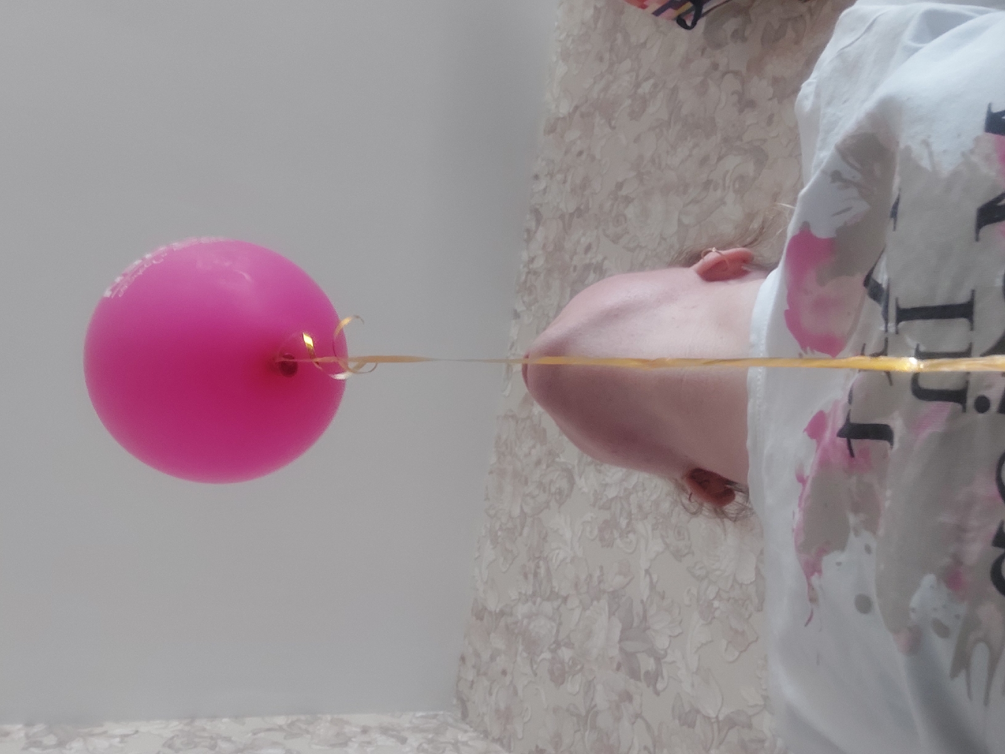 A pink balloon is attached by a gold ribbon to the nose of a woman. The woman is wearing a white t-shirt with black and pink designs. The background consists of a textured wall. The image appears to be taken indoors. There is no information to indicate this photo was taken in the city of Chaikovsky, Russia.