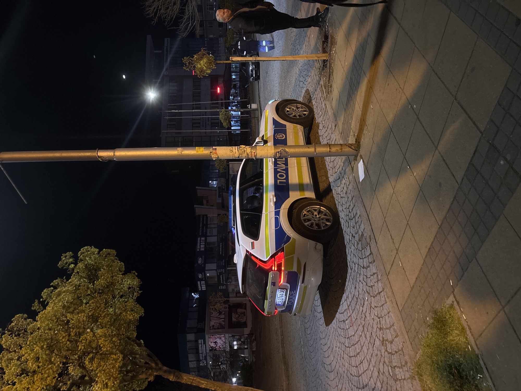 A nighttime scene in Kardzhali, Bulgaria, shows a white and blue police car parked on the sidewalk, partially obstructing a metal street lamp post. A person stands near the front of the car, and a young tree is planted next to the pole. The sidewalk is paved with gray bricks, and the background features buildings, street lights, and other vehicles. The license plate on the police car reads "CB 2744 OB". The word "ПОЛИЦИЯ" (Police) is visible on the side of the vehicle.