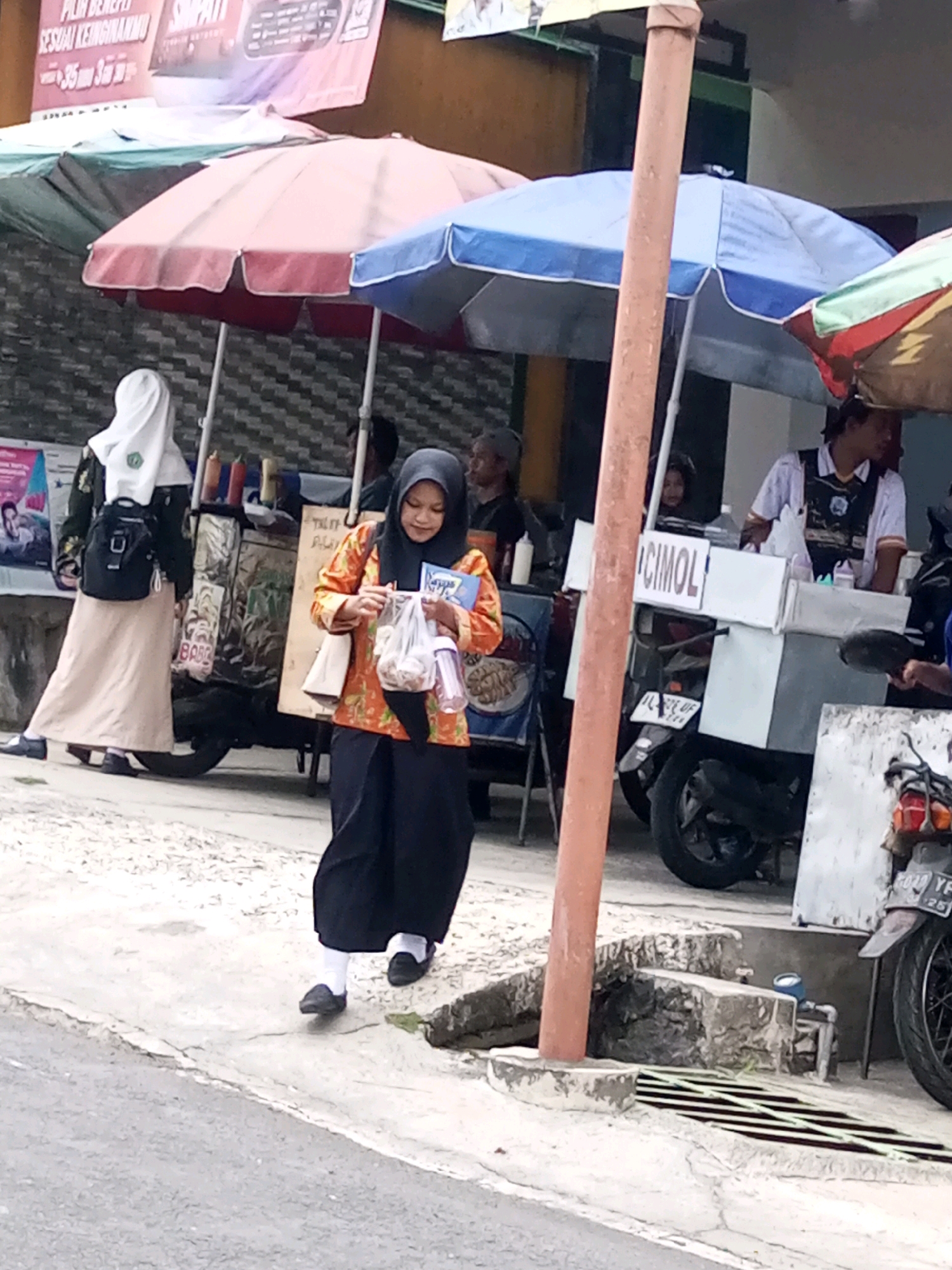In Krasak, Indonesia, an outdoor scene unfolds near a street lined with food stalls and parked motorcycles. A young woman in a black hijab and an orange patterned blouse walks on a concrete path, carrying a plastic bag containing food items and a blue bottle. Behind her, another woman in a white hijab and a long skirt walks away from the camera. Food carts with umbrellas and various merchandise are visible, as is a motorcycle with a license plate. The ground is a mix of asphalt and cracked concrete, with a drainage grate in the foreground. The atmosphere appears to be that of a bustling local marketplace or street food area.