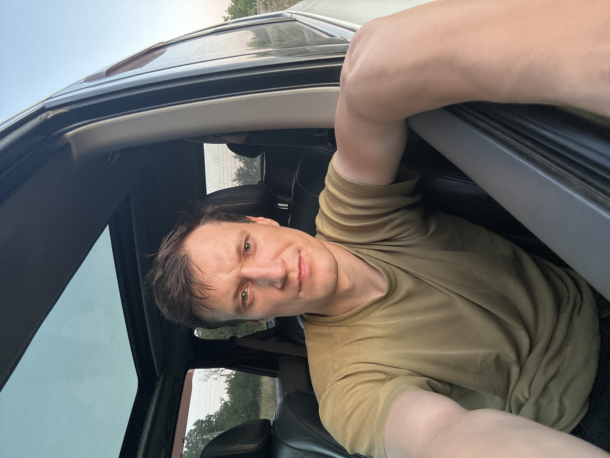 A man with short brown hair and green eyes is sitting in the driver's seat of a car. He is wearing a tan t-shirt and his left arm is resting on the car door. The car has a sunroof, and the windows are open, revealing a landscape of trees and fields under a clear sky. The lighting suggests it is late afternoon or early evening. The image appears to be a selfie taken by the man. The location is identified as Makiv, Ukraine.
