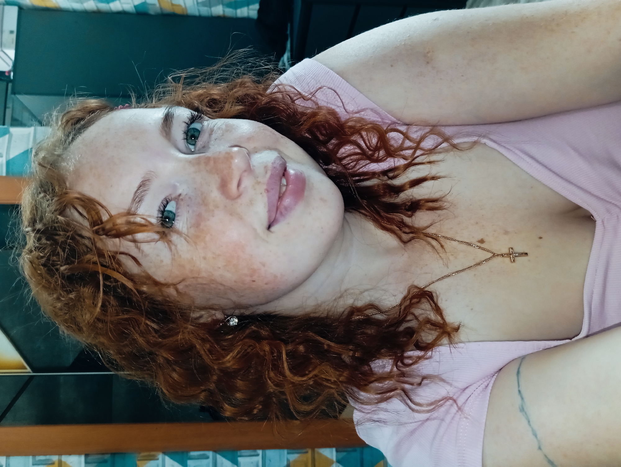 A close-up, angled portrait shows a Caucasian woman with red, curly hair and fair skin with freckles. She has green eyes and is wearing a light pink t-shirt. A gold chain necklace with a small cross pendant is visible around her neck, and she has an earring in her left ear. A blue-green and tan patterned surface is visible at the bottom of the frame. A black surface is in the top background. There is no information to confirm the location to be Yurgamysh, Russia.