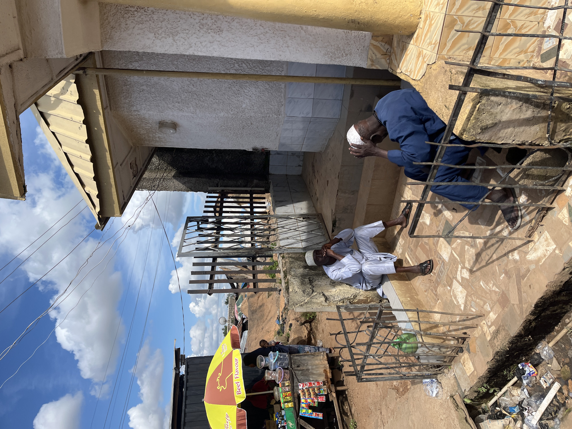 The image depicts an outdoor scene in Gura Top, Nigeria, under a partly cloudy sky. Two men are seated on concrete steps in front of a building. The man on the left, wearing a white cap and light-colored traditional clothing, is seated with his legs crossed and appears to be smoking. The man on the right, wearing a blue traditional outfit and a white cap, has his head in his hands. Adjacent to the building is a stall with a yellow and red umbrella advertising "Beef Flavour." The stall displays various packaged goods and containers. A metal gate is visible behind the men. The ground is uneven and appears to be a mix of dirt and tiled surfaces. A drainage ditch filled with debris is located in the foreground to the left. Overhead, power lines are strung across the sky.