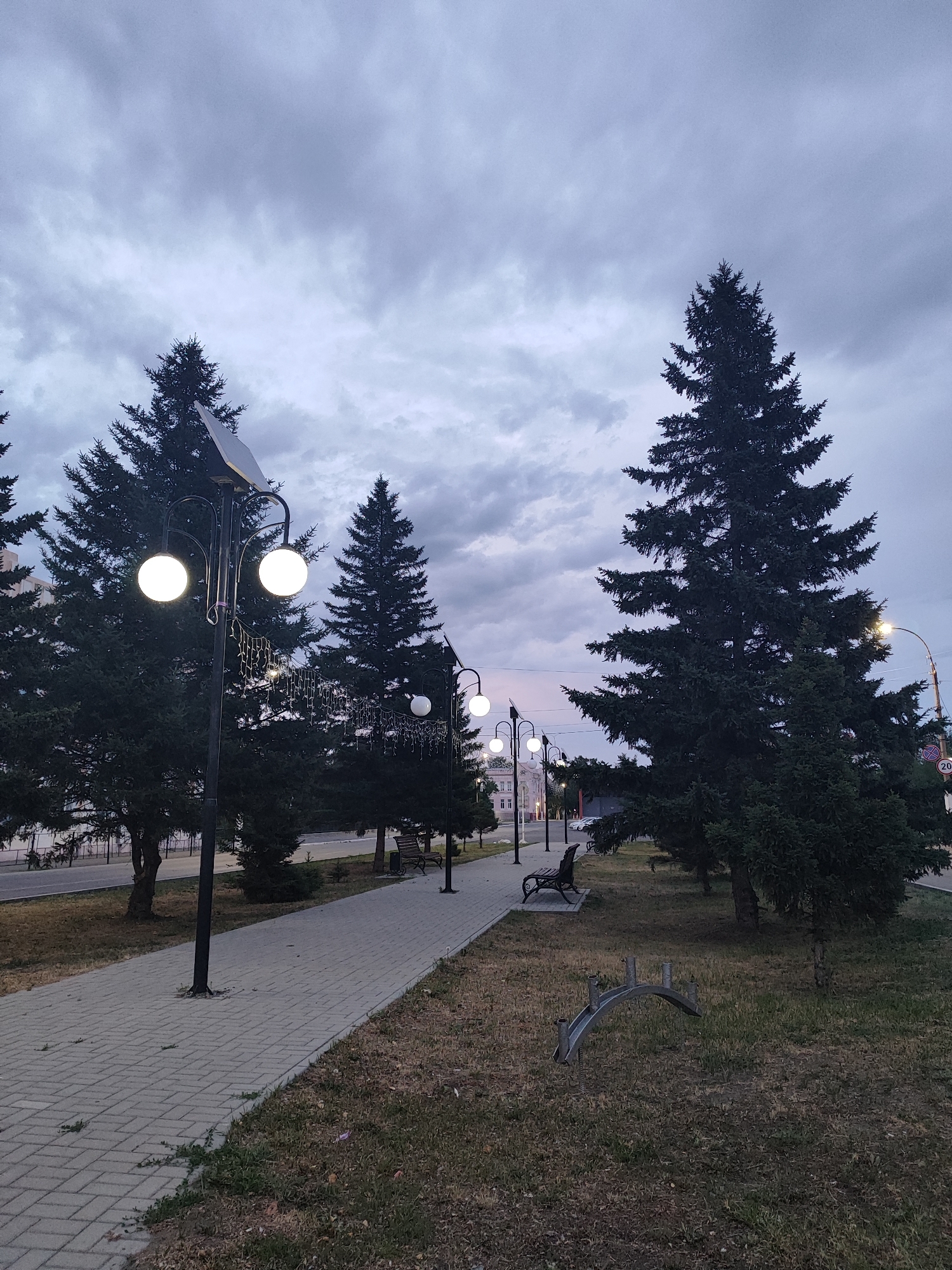 This media file depicts an outdoor scene in Slavgorod, Russia, likely during dusk or twilight. A paved walkway, constructed with grey rectangular pavers, runs diagonally from the left foreground towards the right middle ground. Flanking the walkway is a strip of dry grass interspersed with some sparse green vegetation.
Several large evergreen trees, appearing to be fir or spruce, dominate the landscape. Their dark green foliage contrasts with the muted tones of the sky. A series of lampposts are visible along the walkway. The closest lamppost features two spherical, illuminated lamps and a solar panel at its apex. A string of small, decorative lights hangs from this lamppost, adding a festive touch. Further down the walkway, other lampposts with single illuminated spheres are visible.
In the middle ground, a park bench with an ornate metal design sits on the grassy area. Beyond the trees on the left, glimpses of buildings suggest an urban or suburban setting. The sky is overcast with heavy, grey clouds, indicative of cloudy or impending rain. The overall mood is tranquil and somewhat somber due to the low light conditions and the cloudy sky. No people are visible in the frame. A sign with a circular shape and what appears to be the number "20" in white on a red background is visible on a lamppost in the far right background.