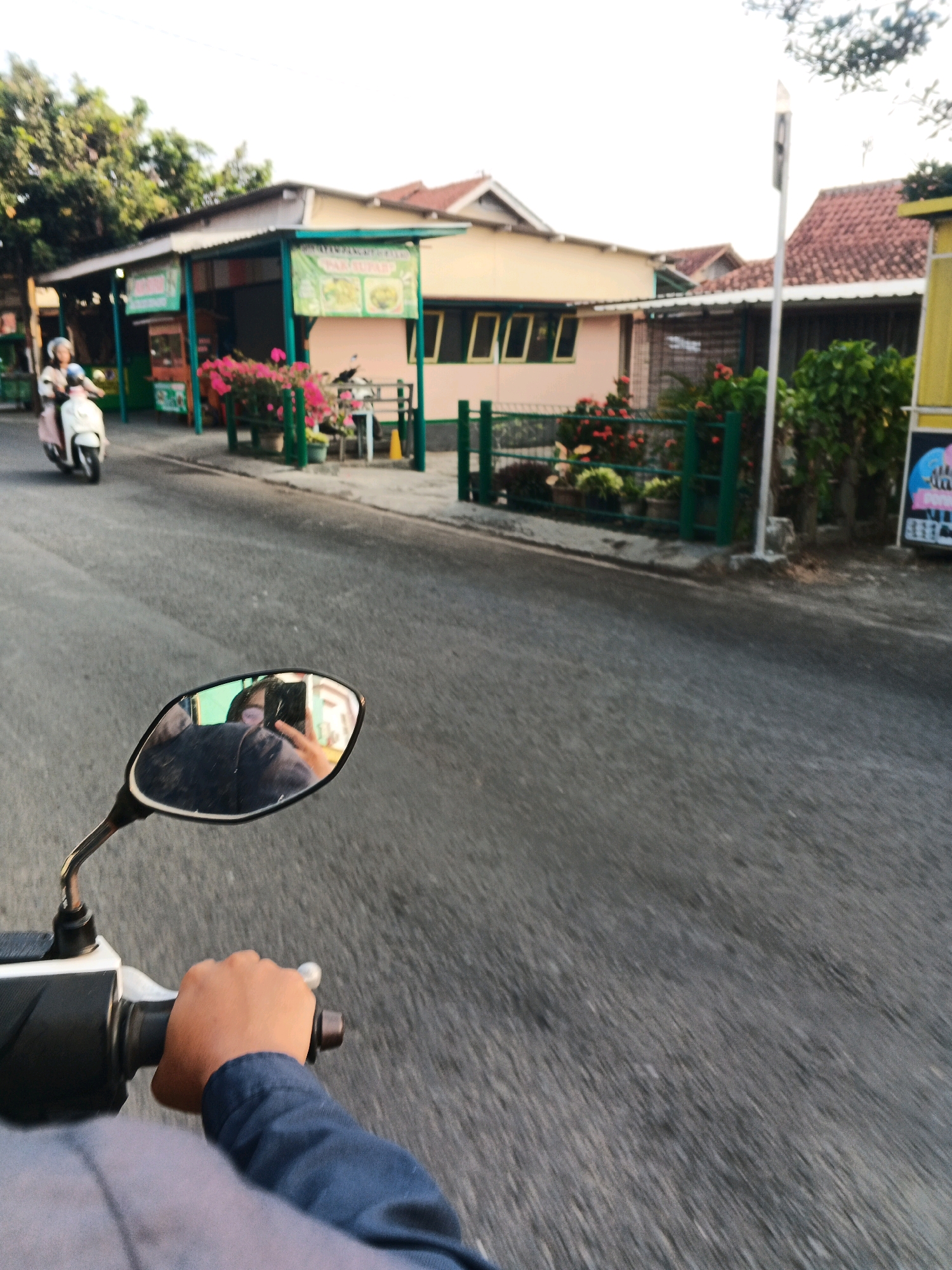The image captures a first-person perspective from a motorcycle in Cilacap, Indonesia. A person's hand, wearing a dark blue long-sleeved shirt, is seen gripping the motorcycle's handlebar. In the left side of the frame, a motorcycle with a white body and pink accents is visible, ridden by a person wearing a helmet. The motorcycle is moving on an asphalt road, with shops and buildings lining the right side. These buildings have light-colored walls, red tile roofs, and green awnings. Several potted plants and flowers are displayed in front of the shops. A reflective mirror of the motorcycle shows a blurred reflection of the rider wearing a mask. The sky is overcast, and trees are present in the background. The scene suggests a typical street view in a commercial area of Cilacap during daytime.