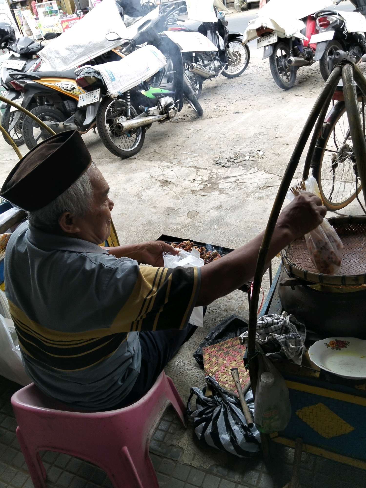 A man, appearing to be of Indonesian ethnicity and elderly, sits on a pink plastic stool in what appears to be a street market. He wears a dark brown cap and a gray and yellow striped shirt. His left hand is holding a transparent plastic bag containing food items, while his right hand is reaching into a black grilling pan filled with what seems to be cooked food, emitting a slight haze. In the background, several motorcycles are parked. One motorcycle, an orange and black model, has a license plate visible that reads "AA 3379 PC." Another motorcycle, a black and white model, is partially obscured. To the right of the man, a woven basket is situated on a wooden stand, next to a white plate with floral designs and a blue and yellow patterned surface. Various bags, including a black and white striped plastic bag, are scattered around the foreground. The ground is paved with concrete and appears somewhat worn. The overall scene suggests a street food vendor's stall in Kemiri Kidul, Indonesia.