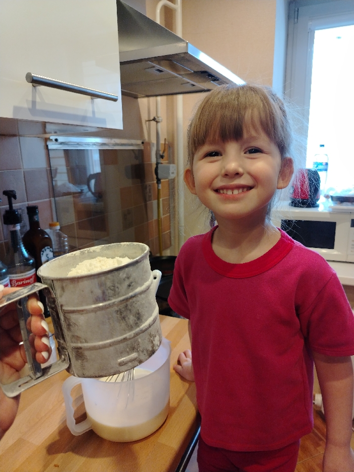 The scene depicts a domestic kitchen in Chaikovsky, Russia, during food preparation. A young Caucasian child, approximately 4-6 years old, with light brown hair and bangs, is positioned on the right, facing forward with a wide smile. The child wears a solid pink short-sleeved t-shirt with a red collar. In the foreground, a left adult hand with white-painted fingernails holds a silver-colored, cylindrical flour sifter filled with white powder over a translucent white plastic measuring jug containing a pale yellow liquid and a metal whisk. The sifter is positioned directly above the jug. The cooking activity takes place on a light-colored wooden or laminate countertop. In the background, a white overhead cabinet with a brushed metal handle is visible on the left, beneath a stainless steel range hood. Further back, light brown tiled walls are present. Various items are on the countertop, including dark glass bottles, one labeled "Bariano." On the far right, a white-framed window allows natural light, and a white microwave oven is visible beneath it. A vertical gas pipe runs along the tiled wall.