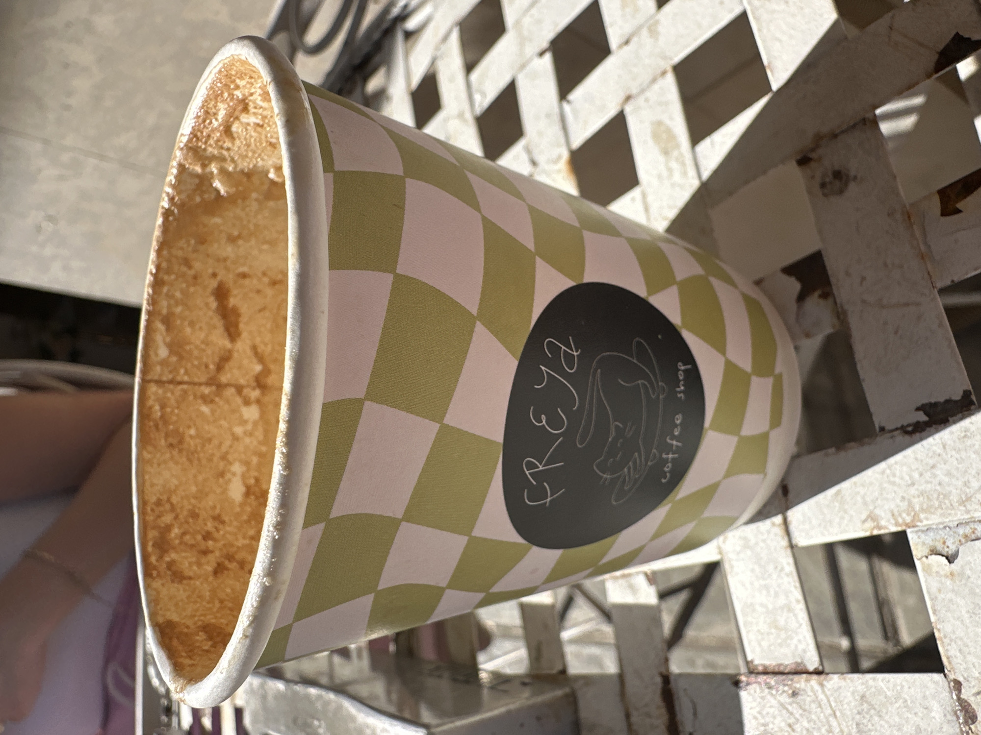 A disposable coffee cup lies on its side on a white, lattice-patterned metal surface, likely an outdoor table or chair. The cup is adorned with a green and light pink checkerboard pattern. A black circular logo on the side displays white, handwritten-style text that reads "FREYA" above "coffee shop," accompanied by a delicate illustration of a relaxed or sleeping cat.
The interior of the cup is mostly empty but shows significant brown residue and stains, suggesting a recently consumed coffee or similar beverage. The rim of the cup also appears slightly soiled. The metal surface beneath the cup exhibits signs of wear, including chipped paint and rust, particularly along its edges and joints.
Strong, well-defined shadows are cast by the lattice pattern onto the surface, indicating bright sunlight and suggesting it is daytime. In the blurry background on the left, an arm with a faint bracelet can be seen. The overall scene suggests an outdoor setting, perhaps a cafe or seating area, in Sofia, Bulgaria.