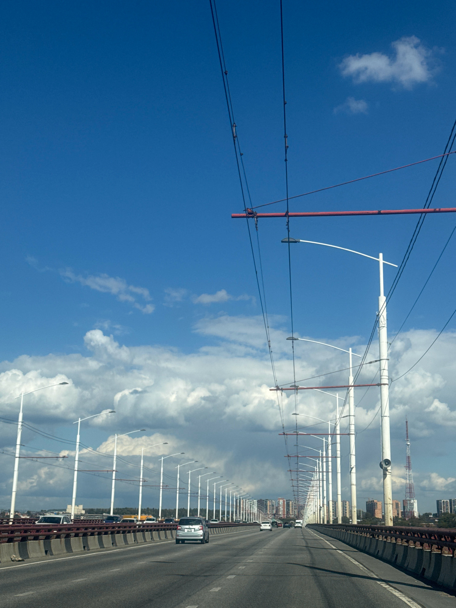 The image depicts a view from a moving vehicle on a multi-lane bridge in Irkutsk, Russia, under a bright blue sky with scattered white clouds. Overhead, a complex network of wires, likely for trolleybuses or trams, stretches across the frame, supported by tall, white poles and red horizontal beams. The bridge has a concrete barrier on the right side and a metal railing on the left, separating the roadway from the side.
The roadway itself is asphalt and appears to be in good condition. Several vehicles, including cars and a silver hatchback, are traveling in the same direction as the viewer. In the distance, a cityscape is visible, with rows of multi-story buildings, some of which appear to be apartment complexes, and a tall, red and white transmission tower. The overall impression is of a functional urban infrastructure scene on a clear day.