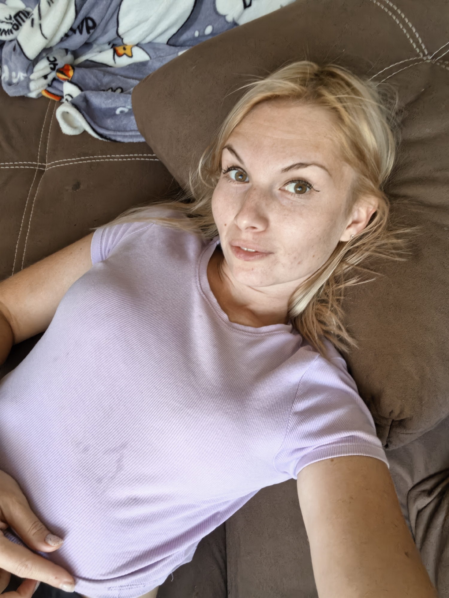 A woman with blonde hair and light skin is lying on a brown couch. She is wearing a light purple t-shirt. A grey blanket with white cartoon penguins on it is partially visible to her left. Her face is tilted upwards and she is looking towards the camera. She has freckles on her face and is wearing black eyeliner.
