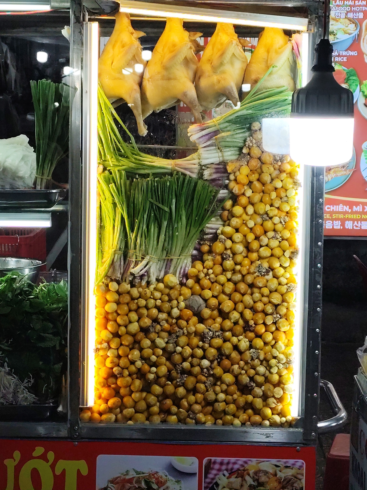 Here's a detailed description of the image:
**Scene:** A brightly lit food stall in Da Lat, Vietnam.
**Subjects/Objects:**
* Four whole cooked chickens are hanging from the top of a glass display case.
* Bunches of scallions and green onions, tied together, are stacked below the chickens.
* A large pile of small, yellow-orange quail eggs fills the bottom portion of the display case.
* On the left side of the food stall, there are containers with fresh herbs and vegetables, including basil.
* A hanging light fixture with a bright LED bulb is visible.
* Signage with Vietnamese and Korean text is partially visible, along with images of food dishes.
**Environment:**
* The setting appears to be an outdoor food vendor area or street market, based on the stall setup.
**Overall Impression:**
The image presents a food stall in Da Lat, Vietnam, showcasing prepared ingredients for dishes. The chickens, various greens, and quail eggs are displayed to attract customers. The signage indicates that the stall offers a variety of dishes, likely including seafood hotpot, stir-fried noodles, and rice dishes.