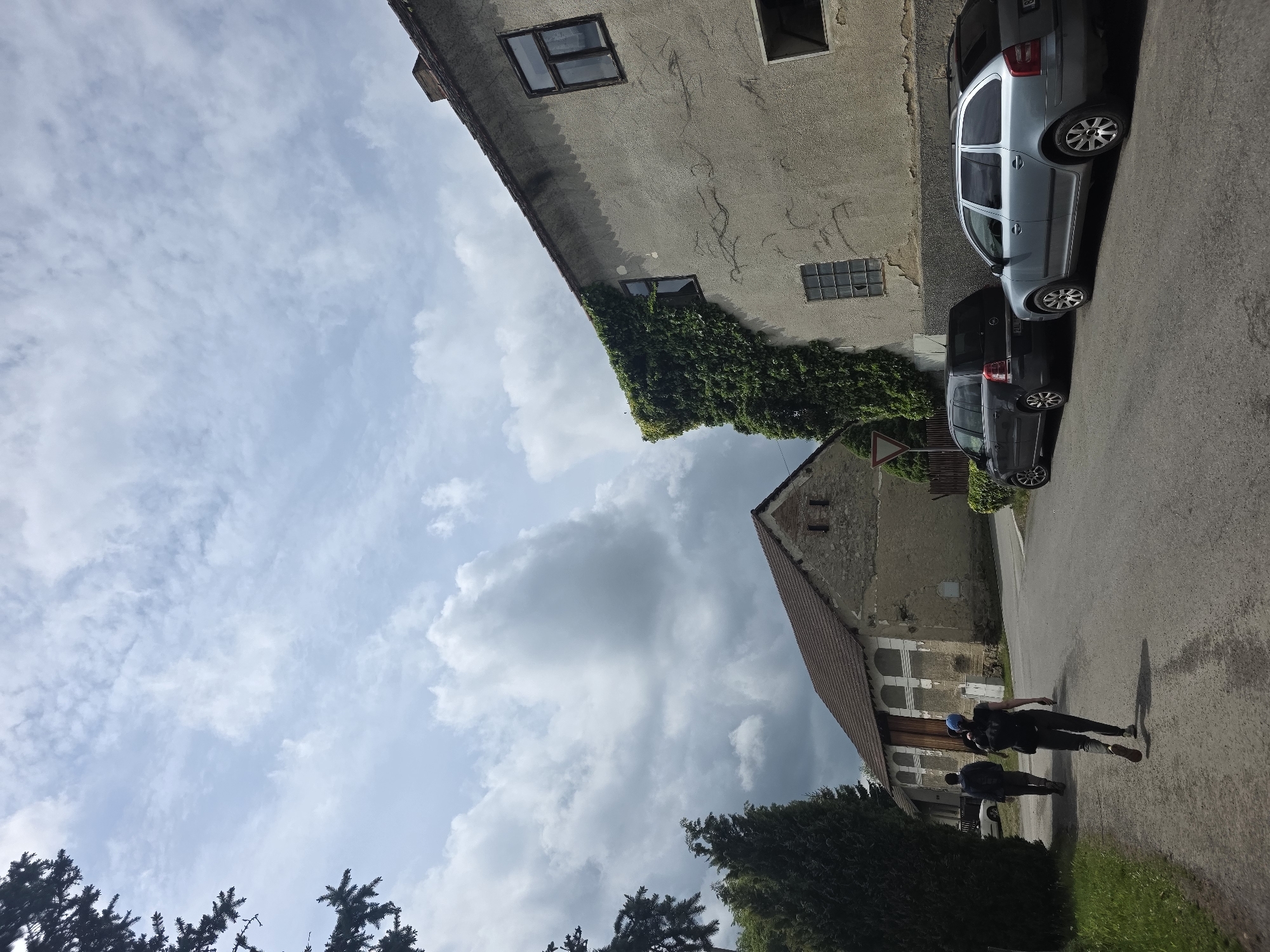The image captures a street scene in an unidentified location. A light gray and a dark gray car are parked on the right side of the street, near a building with ivy growing on its wall. Two people are walking on the right side of the street. The architecture of the buildings suggests a European setting. The sky above is filled with clouds, indicating daylight. Trees are visible in the lower part of the image. The image is rotated.