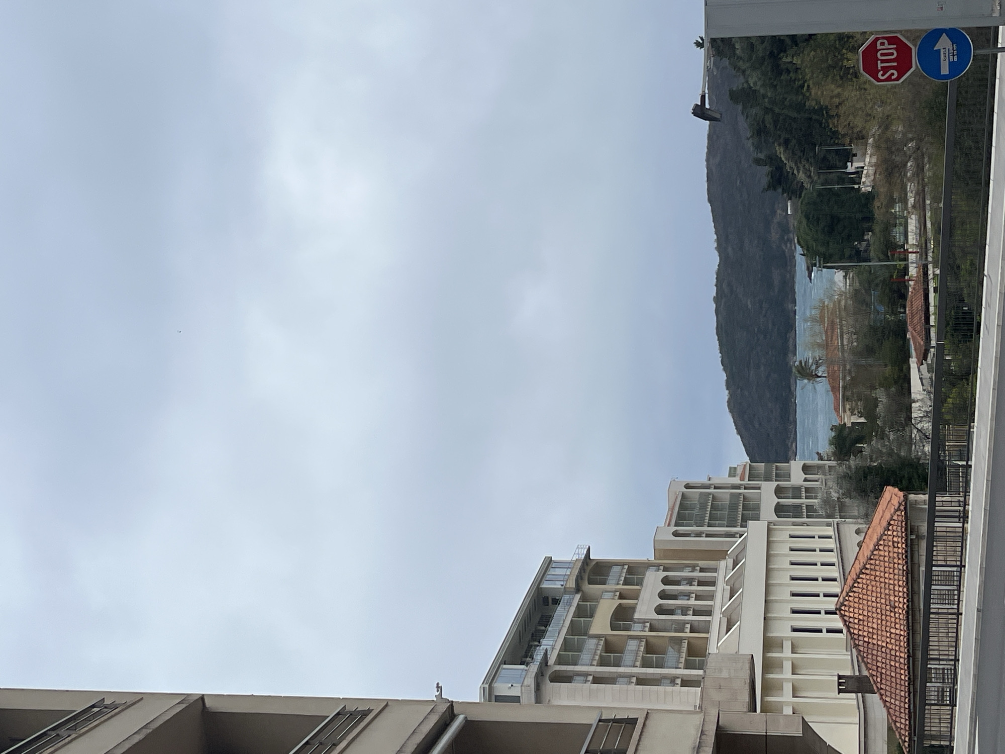The image shows a view in Boreti, Montenegro. In the foreground are buildings. In the middle ground are more buildings, some with balconies. A red tile roof is visible amongst the buildings. In the background is a mountain and a body of water. A stop sign and a one-way street sign are visible. The sky is overcast.