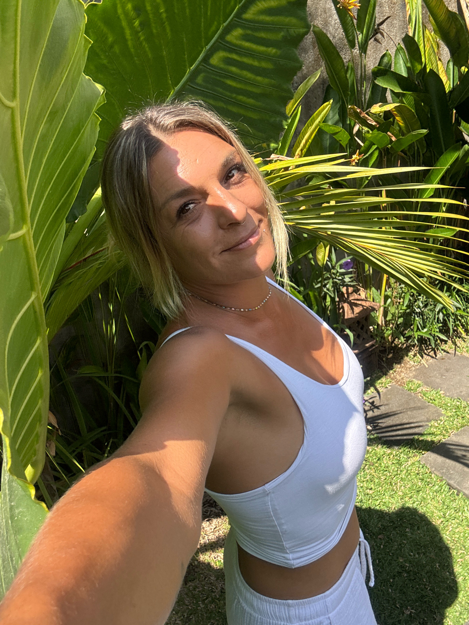 A woman with blonde hair and a light complexion is taking a selfie outdoors. She is wearing a white tank top and white shorts. The background consists of lush green foliage and a stone wall. The lighting suggests it is daytime, likely in a tropical or subtropical climate. Based on the foliage and architectural style, the scene could be located in Ubud District, Indonesia.