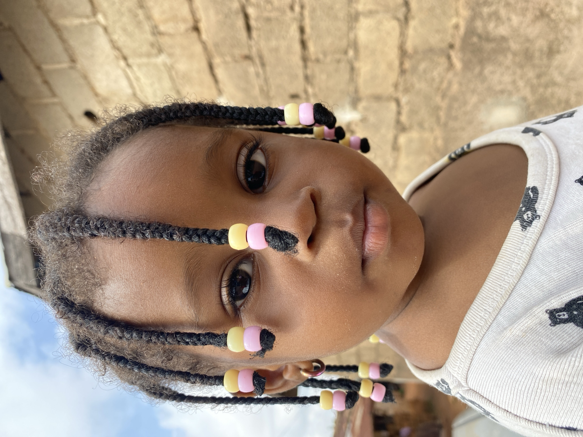 In Bukuru, Nigeria, a close-up portrait captures a young Black child's face from the shoulders up. The child has dark skin and dark, braided hair adorned with alternating yellow and pink beads. The child is wearing a light-colored t-shirt with a repeating pattern of black bear-like figures. The background consists of a textured brick wall and a hint of a cloudy sky. The child's gaze is directed forward, with a neutral expression.