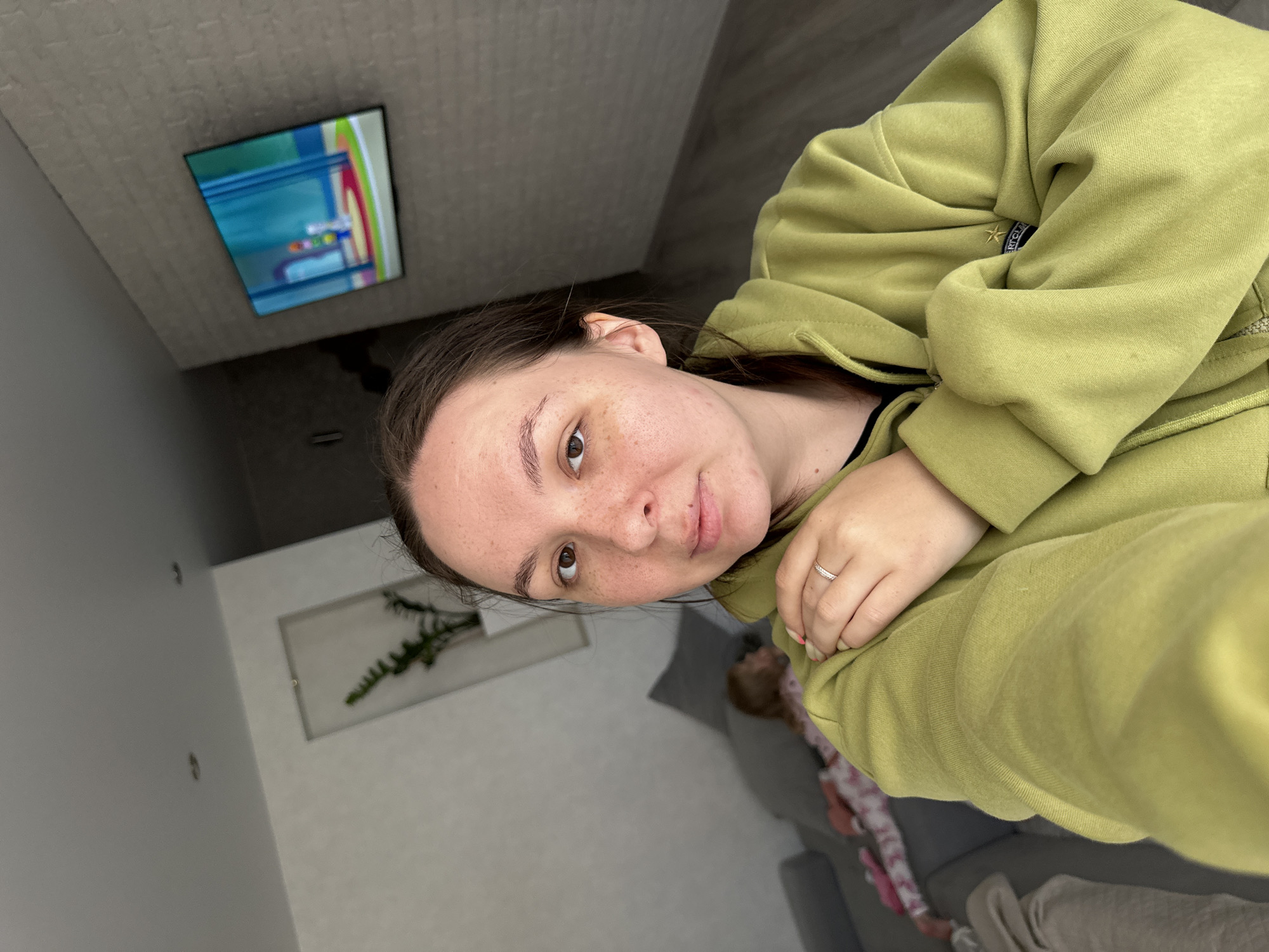 This indoor selfie, taken in Chaikovsky, Russia, features a woman looking directly at the camera. The image is rotated 90 degrees clockwise.
The main subject is a woman with medium-dark brown hair, pulled back from her face. She has fair skin with numerous freckles visible across her nose and cheeks. Her dark brown eyes meet the camera with a neutral expression, possibly a subtle hint of a smile. She wears a light green (lime green) hooded sweatshirt. A silver-colored ring with small stones adorns the ring finger of her left hand, and her nails are painted light pink. On her hoodie, near the neckline, a small golden star emblem is visible, possibly part of a brand logo or design, with a dark blue tag beneath it that has illegible white text.
In the background, a child is partially visible, lying on a grey sofa and wearing pink and white patterned pajamas. The room features light-colored walls and a dark floor. A flat-screen television is mounted on a wall or ceiling, displaying colorful content with blues, greens, yellows, and reds. Also visible is a framed picture or mirror on a wall, containing a green plant that resembles a fern.
The overall impression is of a relaxed, casual moment inside a home, likely during the daytime given the ambient lighting. No legible text is visible in the image.