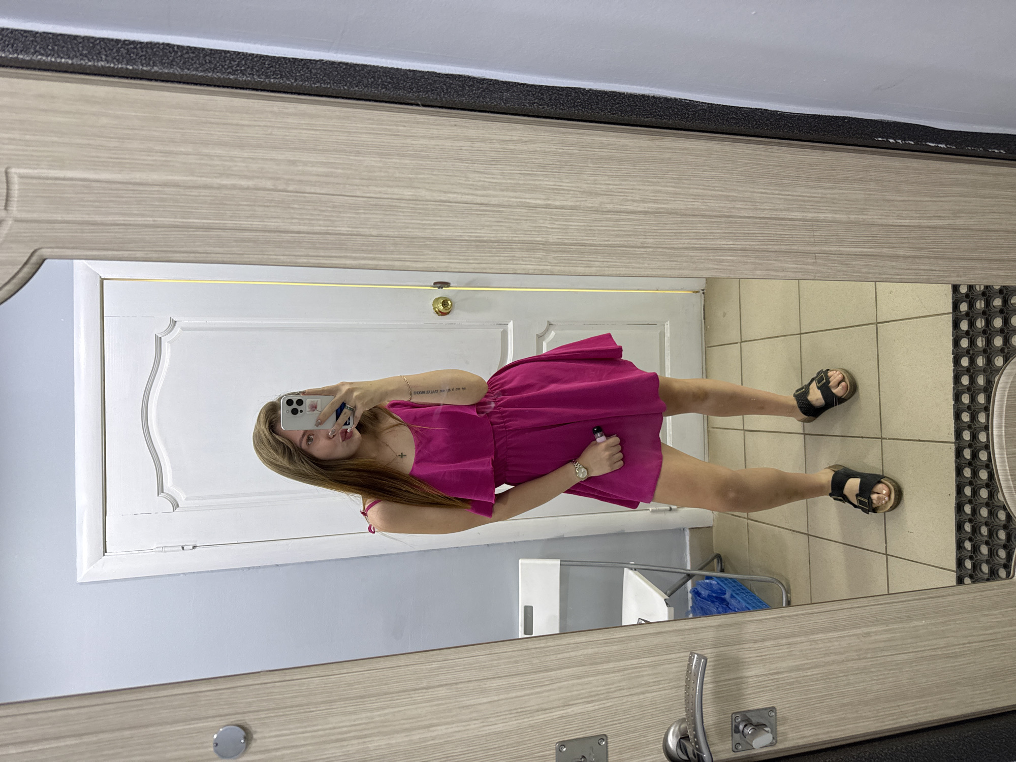 A mirror reflects a woman taking a selfie in what appears to be a bathroom. She's wearing a magenta dress and sandals. A white door is in the background. The location might be inferred as Chaikovsky, Russia, based on external information, but the image itself doesn't provide explicit evidence of this.