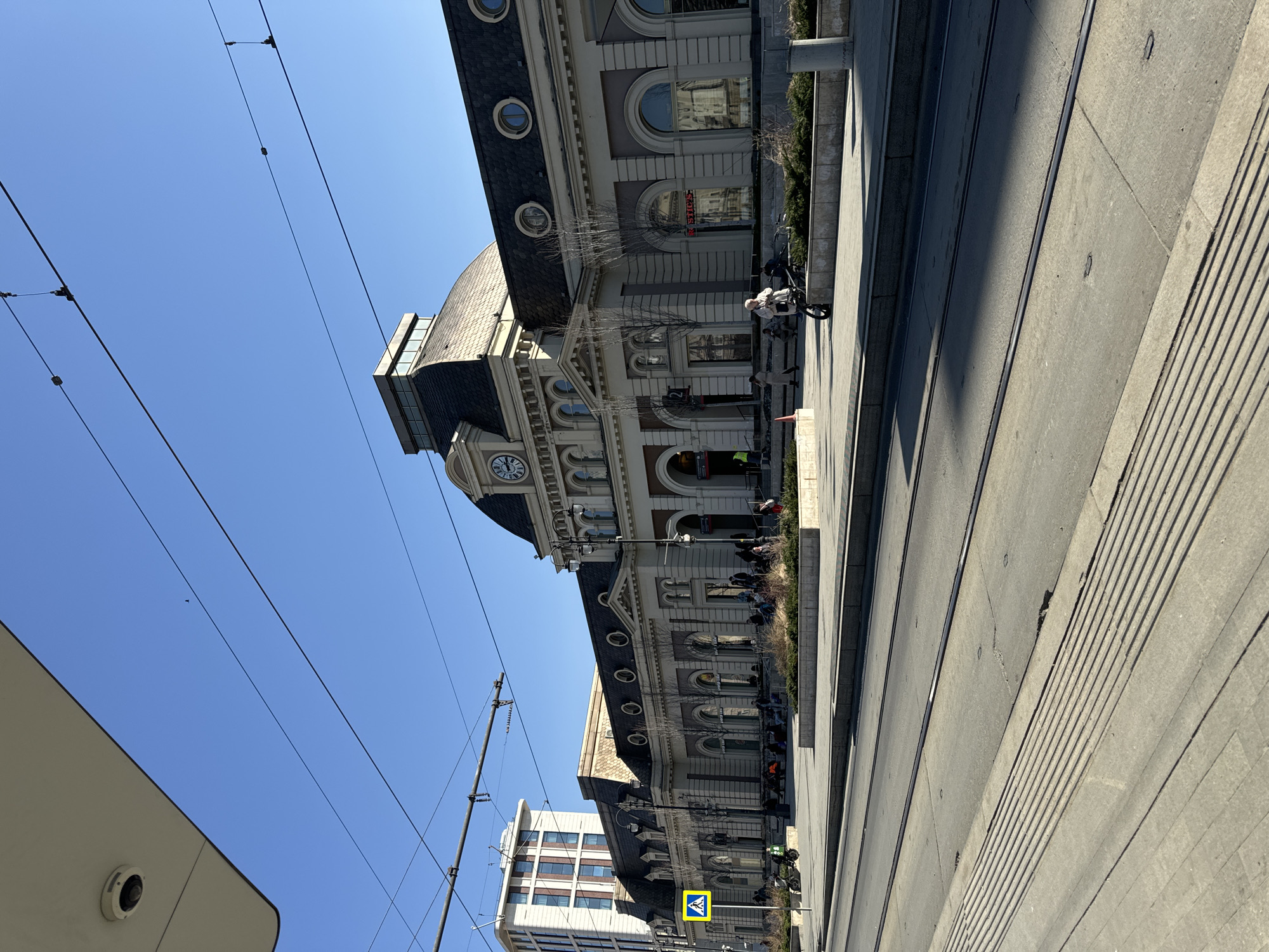 The image depicts a street scene in Moscow, Russia, featuring the Kazansky railway station. The building is a large, ornate structure with multiple windows, a clock tower, and a dark gray roof. A blue crosswalk sign is visible on a white rectangular pole. A modern building with white facade is also visible in the background. Overhead power lines are strung across the clear blue sky. People are present on the sidewalk, some sitting and others walking. The foreground consists of a wide, gray concrete surface with tram tracks. The perspective of the photo is slightly tilted.