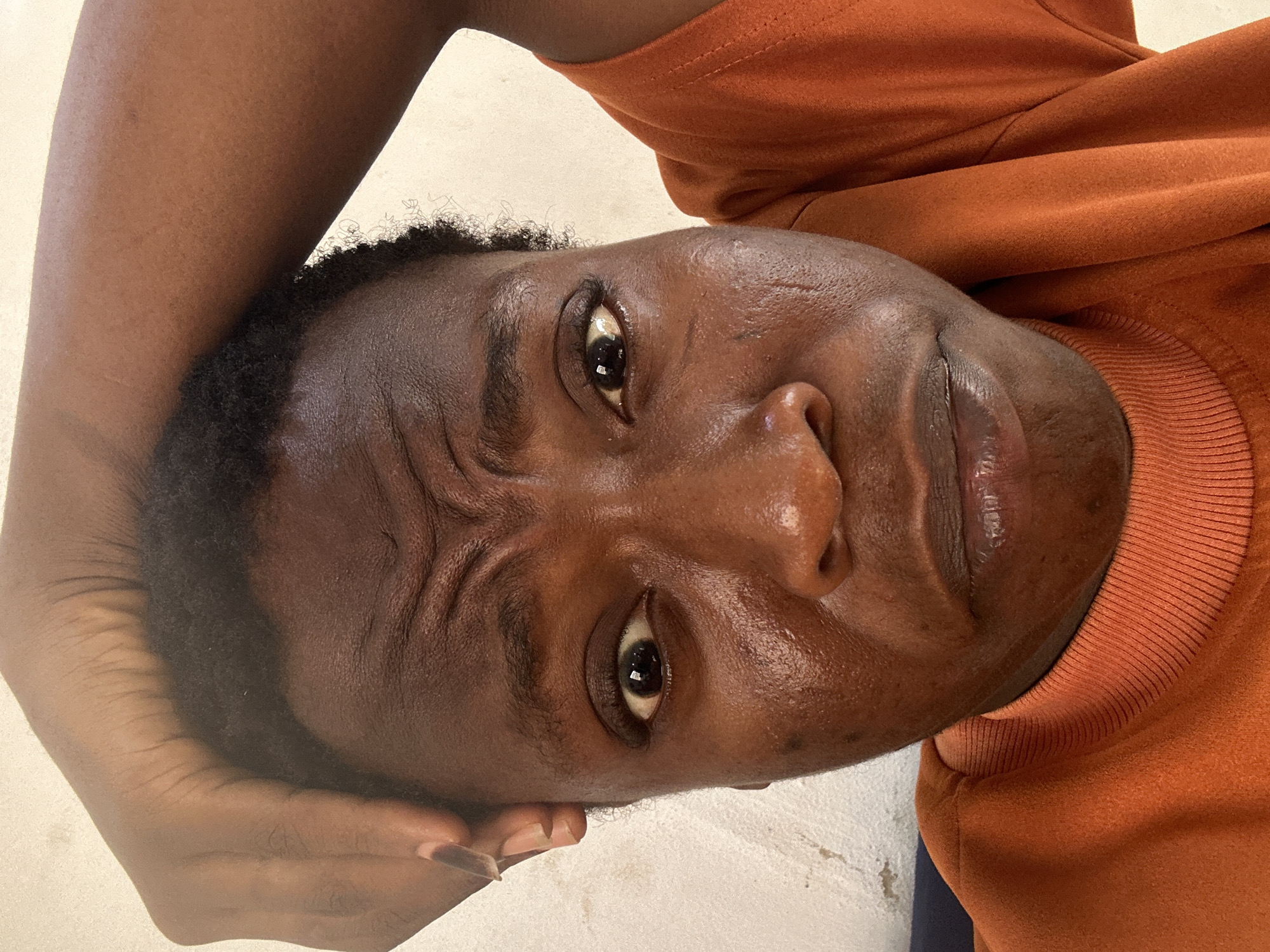 A medium, slightly rotated close-up shot shows a dark-skinned person with short, dark hair wearing a rust-colored sweater. Their face is positioned with their head resting on their hand. There are noticeable creases on the person's forehead. The background appears to be a light-colored surface. The location is Unknown, Unknown.