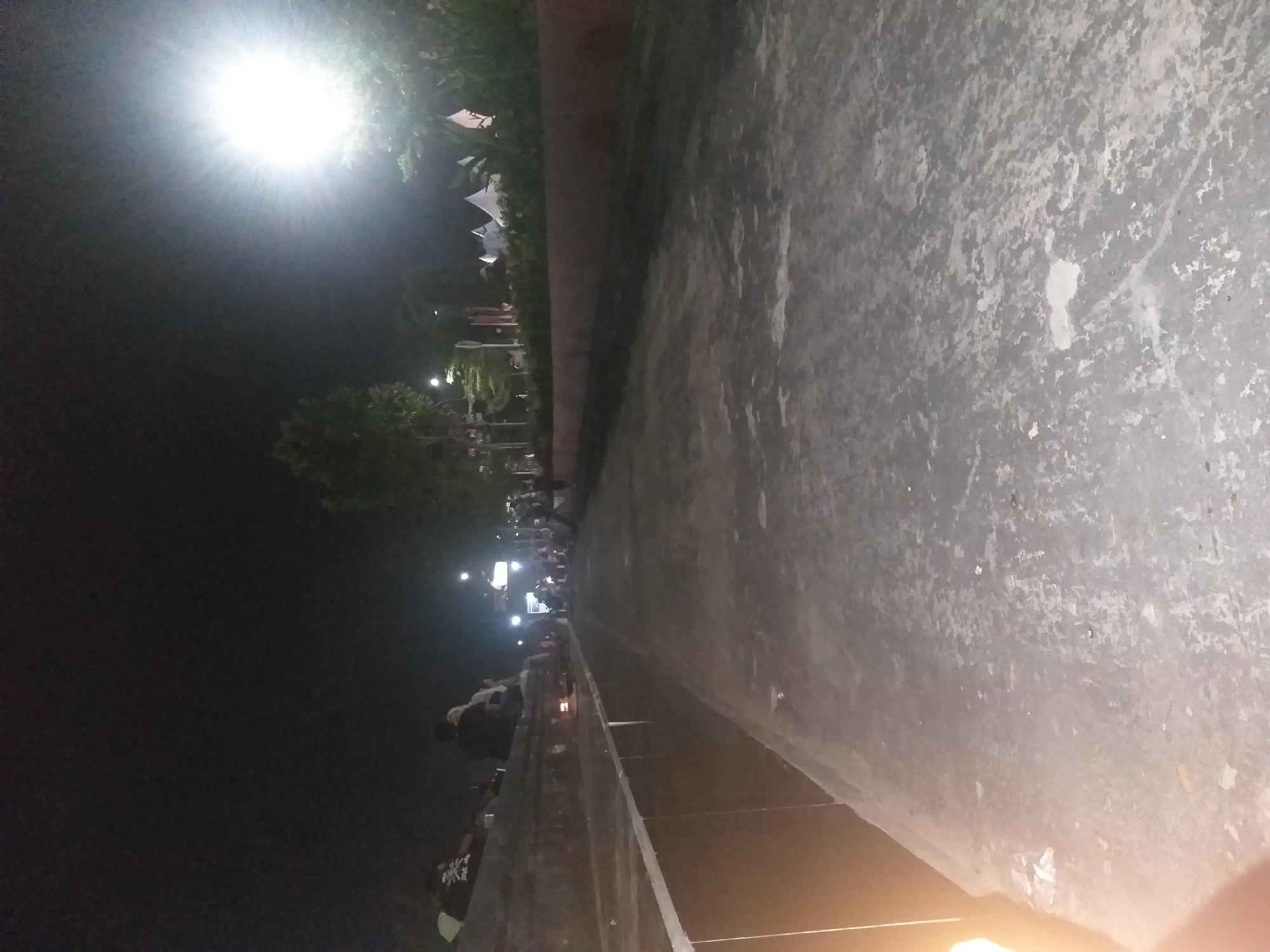 A wide, empty paved walkway runs diagonally across the frame from the lower left to the upper right, illuminated by a bright light source in the upper right. The walkway is textured and appears to be concrete with patches of lighter discoloration. To the left of the walkway is a low, dark wall with a white trim, upon which several people are seated, facing away from the camera. Beyond this wall and in the background, there are trees and foliage, as well as several white tents. A street light is visible in the distance towards the upper left. The scene is set at night, with the sky being dark and featureless.
The location is the waterfront of the city of Dumai, Indonesia, specifically the area known as Pantai Palette. The time is night.