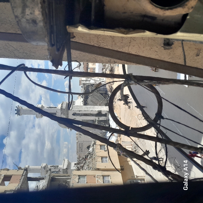 The image displays an elevated view of a heavily damaged urban landscape in Khan Yunis, Palestinian Territory, under a partially cloudy blue sky. The immediate foreground is obscured by dark, twisted metalwork, likely from a damaged balcony or window frame, including a prominent circular element. To the far right, a vertical structural element and a damaged fixture further frame the scene.
In the background, a large mosque with two white minarets is visible. The mosque's main structure exhibits significant damage, with portions of its roof and walls collapsed into rubble. Surrounding buildings also show severe destruction, featuring collapsed sections, exposed interiors, and twisted rebar. Extensive piles of debris and rubble are scattered across an open, sandy area in the midground, situated between the damaged structures and a paved road.
On the paved road and within the adjacent rubble-strewn area, several individuals are present. One person is seen with a dark animal, possibly a donkey or horse, near the center. Other figures walk along the road and through the debris. A small, wheeled cart is visible further down the street to the right. At the bottom left, a person partially clad in red is visible. The entire scene conveys widespread structural devastation.
