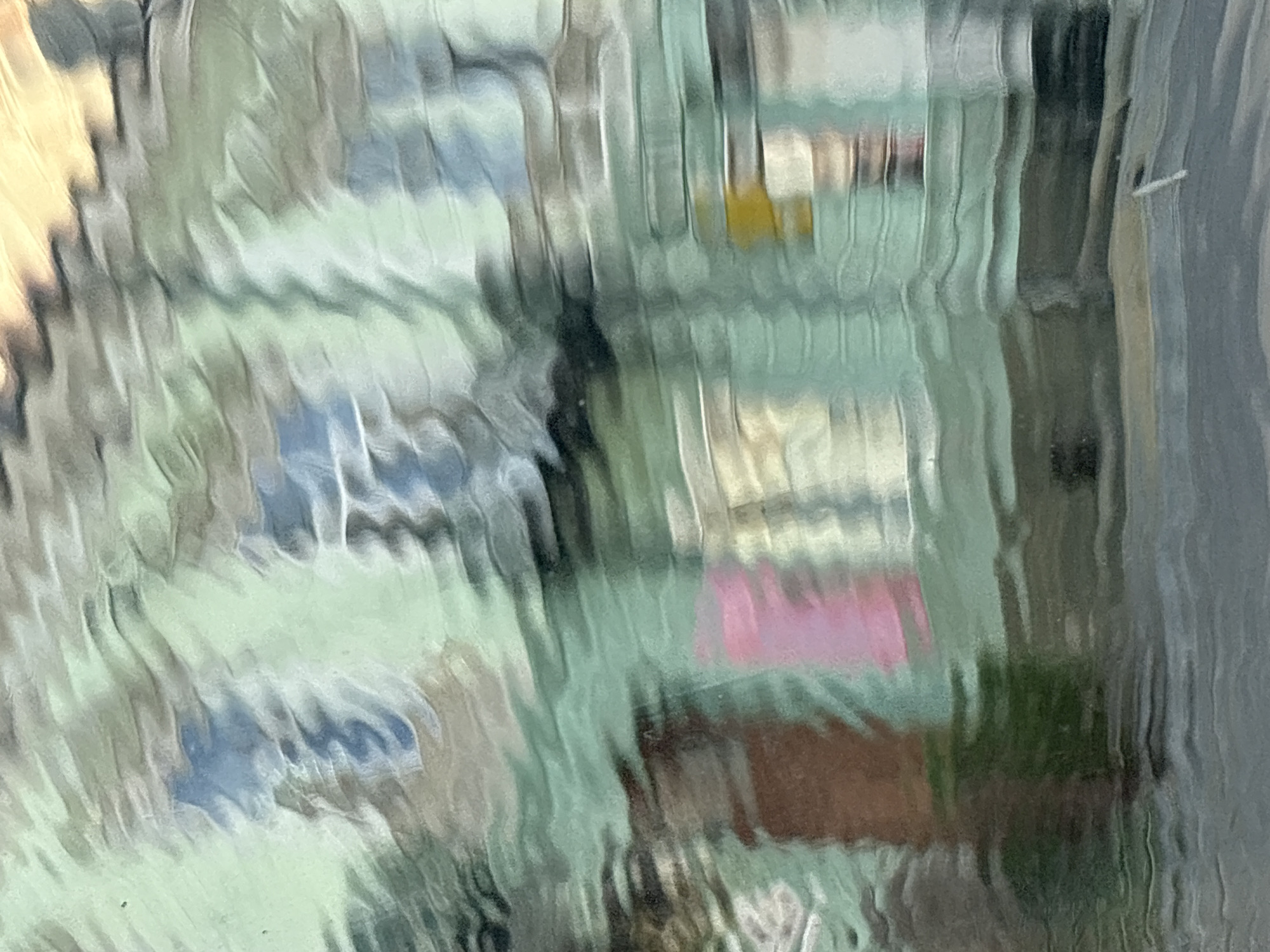 The image depicts an abstract, distorted view of a building facade, possibly viewed through a textured or wet surface. The colors are muted and blended, with dominant shades of light green, blue, grey, and hints of yellow and brown. The distortion creates a wave-like effect across the entire frame, blurring distinct features and suggesting movement or a fluid medium. There are suggestions of windows, doors, and architectural elements, but they are heavily obscured by the visual texture. The specific location in Stryi, Ukraine, is not discernible from the image itself, as the abstract nature of the scene does not provide recognizable landmarks or contextual clues. The image appears to capture a reflection or a view through a material that significantly alters the perception of the underlying subject.