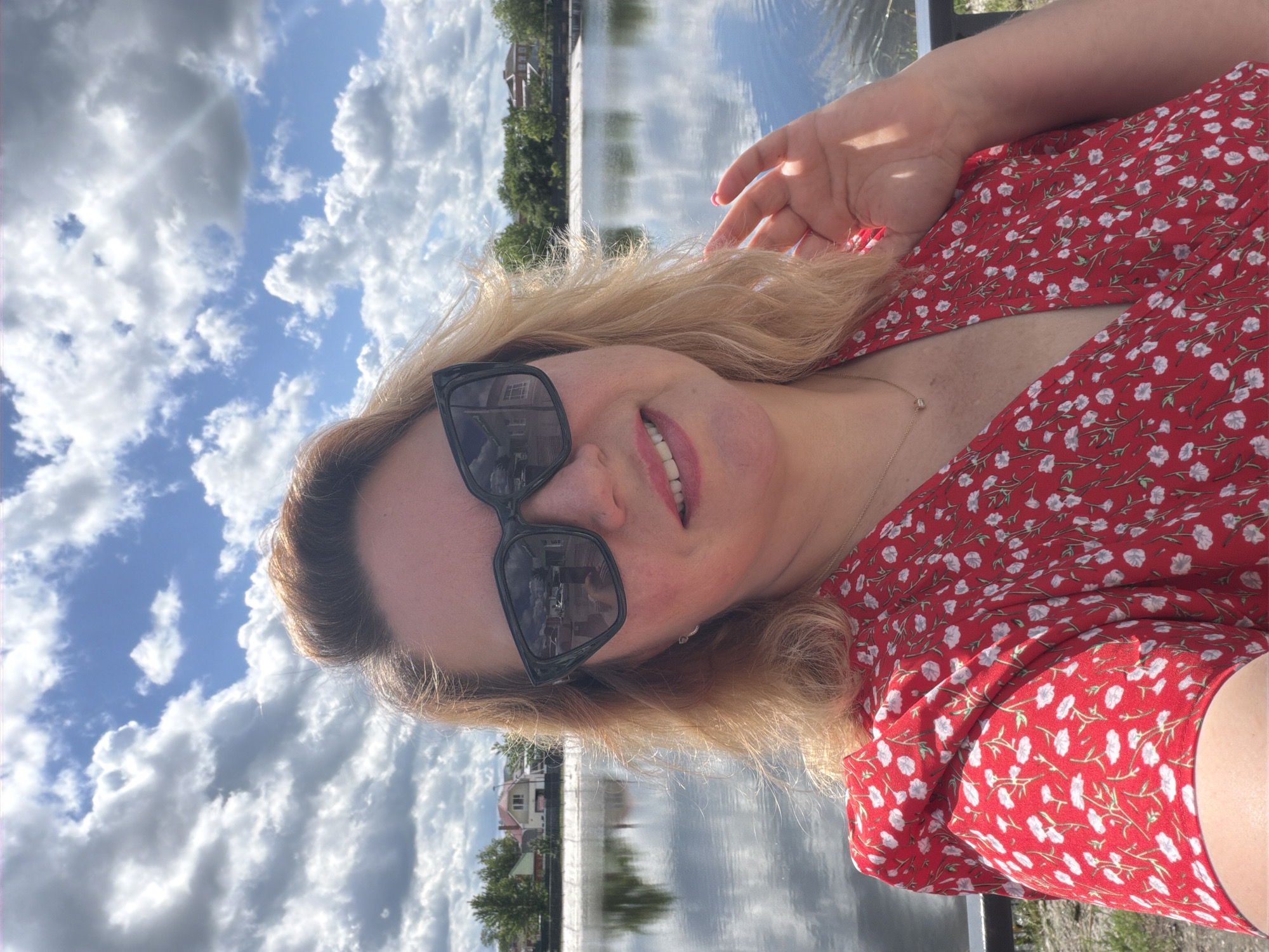 A woman with blonde hair and sunglasses is smiling in a selfie. She is wearing a red dress with a white floral pattern. In the background is a body of water reflecting the sky. The scene appears to be in Leningradskaya, Russia, as evidenced by the Russian architecture visible in the background. The woman has her hand raised slightly in the air. The weather appears sunny with scattered clouds.