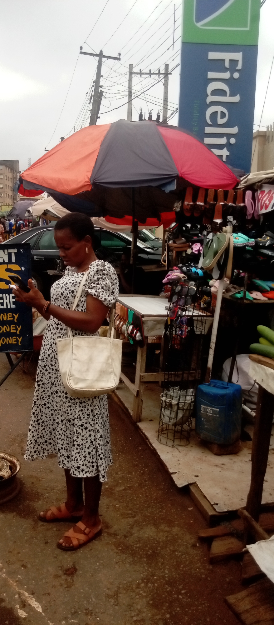 A woman in a white and black patterned dress and brown sandals stands on a street looking at her phone. She has a white shoulder bag. Behind her is a market stall with an umbrella. Shoes and sunglasses are on display at the stall. A sign for Fidelity Bank is visible in the background. Cars are parked nearby. The environment appears to be a busy street scene in an urban area.