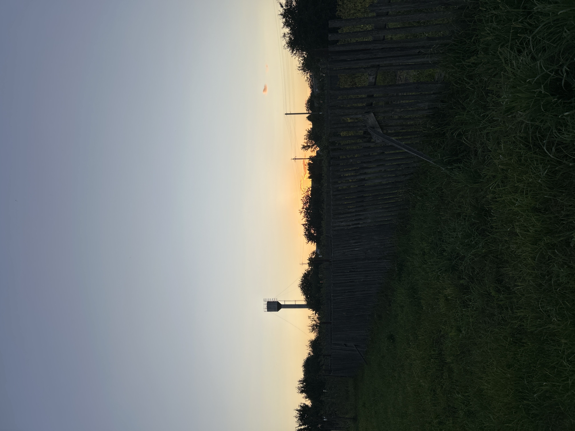 The image depicts a rural landscape at dusk, likely in the Яромінскі сельскі Савет, Belarus. A weathered wooden fence runs diagonally across the frame, separating a field of tall, dark green grass in the foreground from a line of trees and bushes in the midground. In the distance, a water tower stands silhouetted against the pale orange and blue gradient of the sunset sky. A few scattered clouds are visible, one tinged with pink. Several utility poles with wires are also present. The overall impression is one of a quiet, late evening in a rural setting. There are no people or animals visible in the image.