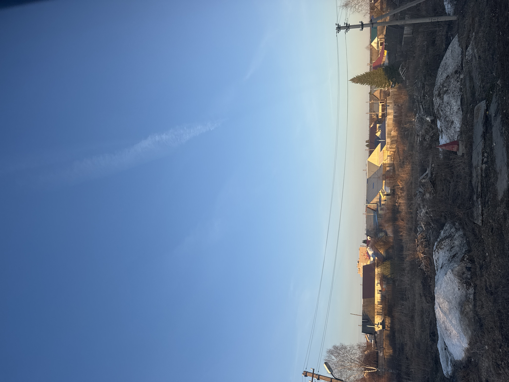 The image captures a serene outdoor scene, likely in Oktyabrsky, Russia, characterized by a vast, clear blue sky and a residential landscape bathed in low sunlight. The sky dominates the upper portion, displaying a gradient from a deep blue at the top to a lighter, more yellowish hue near the horizon, indicative of either early morning or late afternoon. Faint contrails or thin clouds are subtly visible across the blue expanse.
The lower section of the image reveals a semi-rural or suburban setting. The foreground ground cover consists of dry, brown vegetation, dirt, and significant patches of dirty, melting snow or ice, suggesting a late winter or early spring season. A large, irregularly shaped snowdrift occupies the right foreground.
In the midground, a collection of single and two-story houses with varied roof colors (red, yellow, blue) line the horizon. Bare deciduous trees are interspersed among the homes, alongside a distinct, tall evergreen coniferous tree. Utility poles with power lines stretch across the scene, a common feature in such areas. One utility pole in the foreground shows some faded, indiscernible markings. No people or discernible activities are taking place; the scene is static and quiet. The warm, golden light on the buildings and trees suggests the soft illumination of a rising or setting sun.