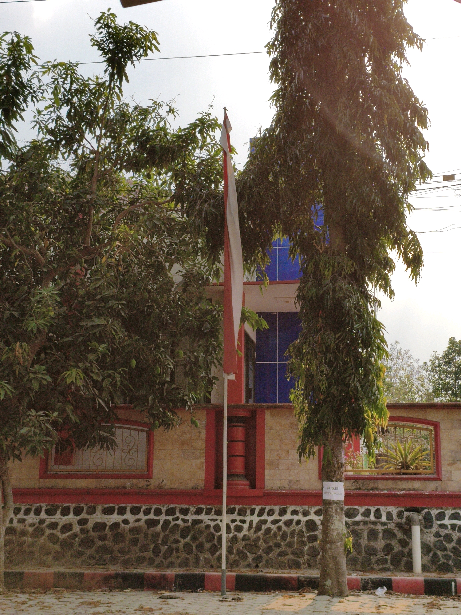 In the city of Puhpelem, Indonesia, a flag pole stands tall, adorned with the Indonesian flag, a symbol of national pride. The flag's vibrant red and white stripes wave gently against the backdrop of a clear sky, hinting at a bright, sunny day. The flag pole itself is rooted in a bed of gravel, nestled beside a paved walkway.
To the left of the flag pole, a lush green tree provides a natural canopy, its branches reaching towards the sky. Its leaves, a myriad of green hues, create a sense of life and vitality. In the distance, a building with a striking blue facade stands tall, its windows reflecting the sunlight. A stone wall, adorned with red accents, separates the building from the foreground, adding a touch of architectural interest.
To the right of the flag pole, another tree, its trunk weathered and rough, stands sentinel. A white sign, attached to its trunk, bears the word "PARKIR," suggesting a designated parking area nearby. The ground beneath the tree is scattered with fallen leaves, a subtle reminder of the changing seasons. The scene is one of serene beauty, a harmonious blend of nature and human creation.