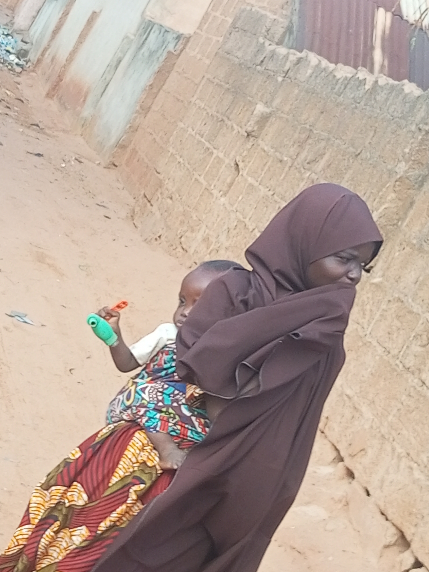 This image depicts two people in an outdoor setting. The primary subject is a woman wearing a dark purple hijab and a matching, flowing garment. She is seen from the side, with her head turned slightly to her right, and her right arm is partially obscuring her face. A young child is strapped to her back, partially visible. The child is wearing a white shirt and a colorful, patterned fabric that is wrapped around them. The child is holding a small green object with a red attachment in their left hand.
The background consists of a dirt path and a wall constructed from rough, rectangular bricks. Above the brick wall, a corrugated metal structure is visible. The scene appears to be in a dusty, possibly arid environment, suggested by the texture of the ground and the muted tones of the walls. The lighting suggests it is daytime, possibly morning or afternoon. There are no visible signs of text in the image. The overall impression is one of everyday life, with a woman carrying a child through a residential or village area. The woman's posture and the way she is holding her arm might suggest a moment of rest or shyness.