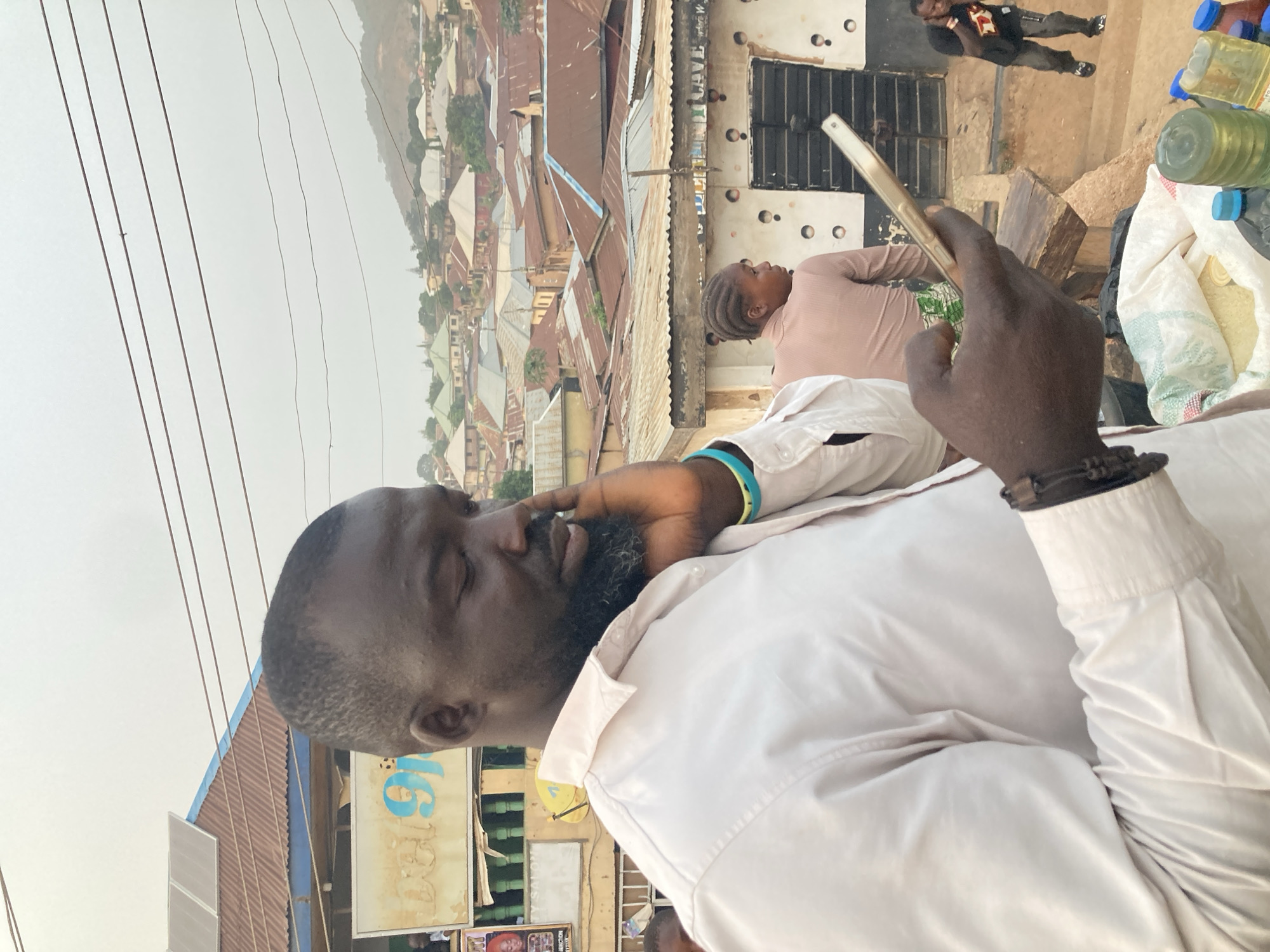 In an outdoor scene set in Gwong, Nigeria, a dark-skinned man with a beard and short hair is prominently featured in the foreground, wearing a light-colored collared shirt. He holds a light-colored smartphone in his right hand and appears to be looking at its screen. A dark bracelet adorns his left wrist, and a light blue and green band is visible on his right arm.
In the mid-ground, a woman with braided hair, dressed in a light pink long-sleeved top, stands near a building. This building has a dark facade adorned with circular patterns. On the ground nearby, several plastic bottles, some containing a yellowish liquid, and a white sack are visible. Further in the background, another person wearing a black t-shirt and jeans can be seen near a doorway.
The background reveals a densely built-up area characterized by numerous buildings with varied corrugated metal roofs in shades of brown, grey, and blue. A hazy mountain range is visible in the distance under an overcast or hazy sky. Multiple power lines traverse the upper portion of the frame. On a rooftop to the left, solar panels are installed.
The general impression is that of a bustling, semi-urban environment. The lighting suggests a midday or late morning/early afternoon, given the soft, diffused quality of light. Partially visible text includes "CAVE" above the woman and another sign with light blue text that is mostly unreadable.
