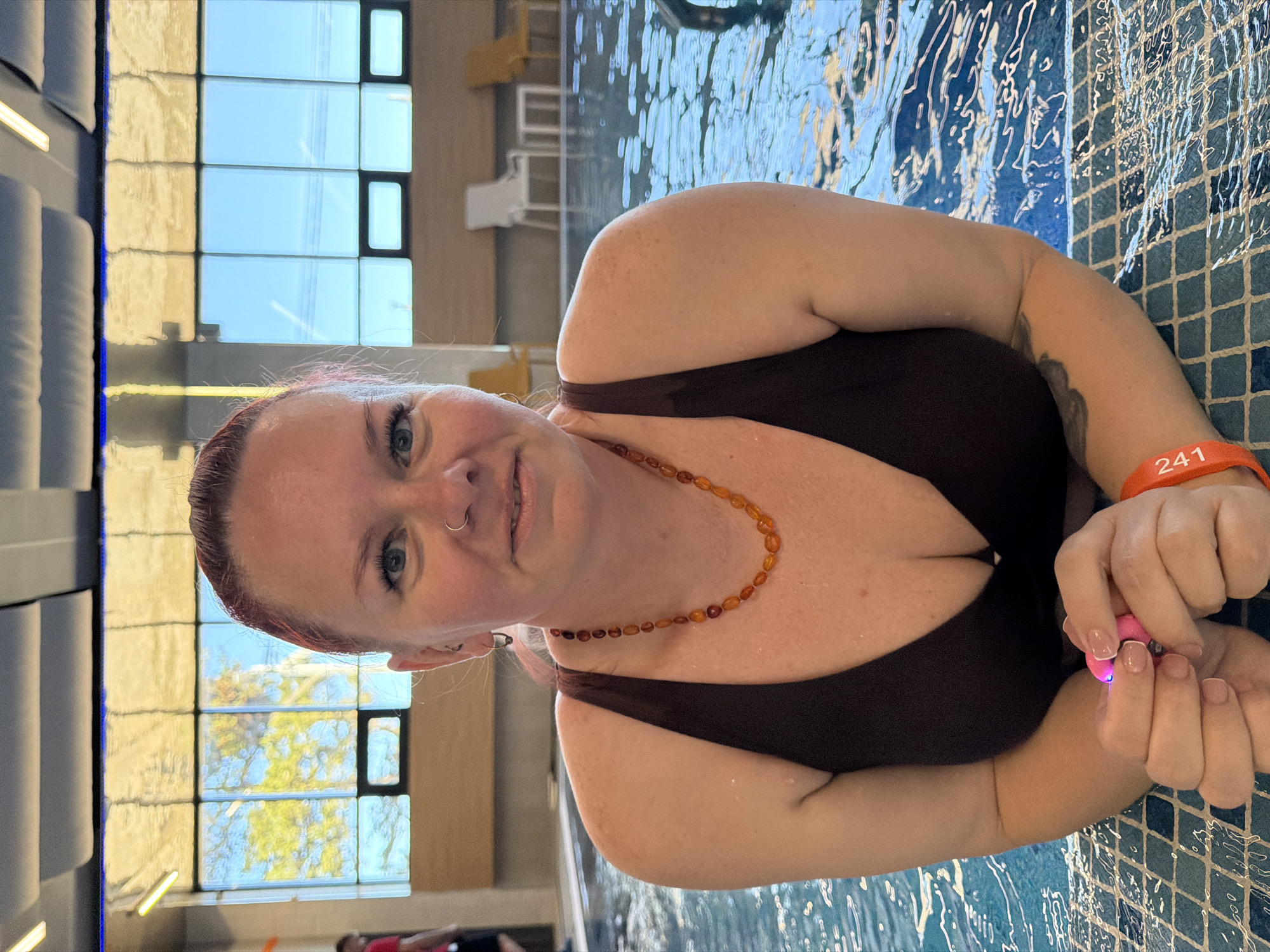 A woman in a dark brown swimsuit is in a swimming pool with blue mosaic tiles. She wears a necklace of amber-colored beads, a nose ring, and an orange wristband with the number "241" on her right wrist. She is holding a small pink object in her hands. Behind her are large windows with reflections of trees and a clear blue sky. The location is inferred to be in Votkinsk, Russia, though the context of the scene suggests an indoor swimming pool facility. There are several chairs and loungers out of focus and in the background.