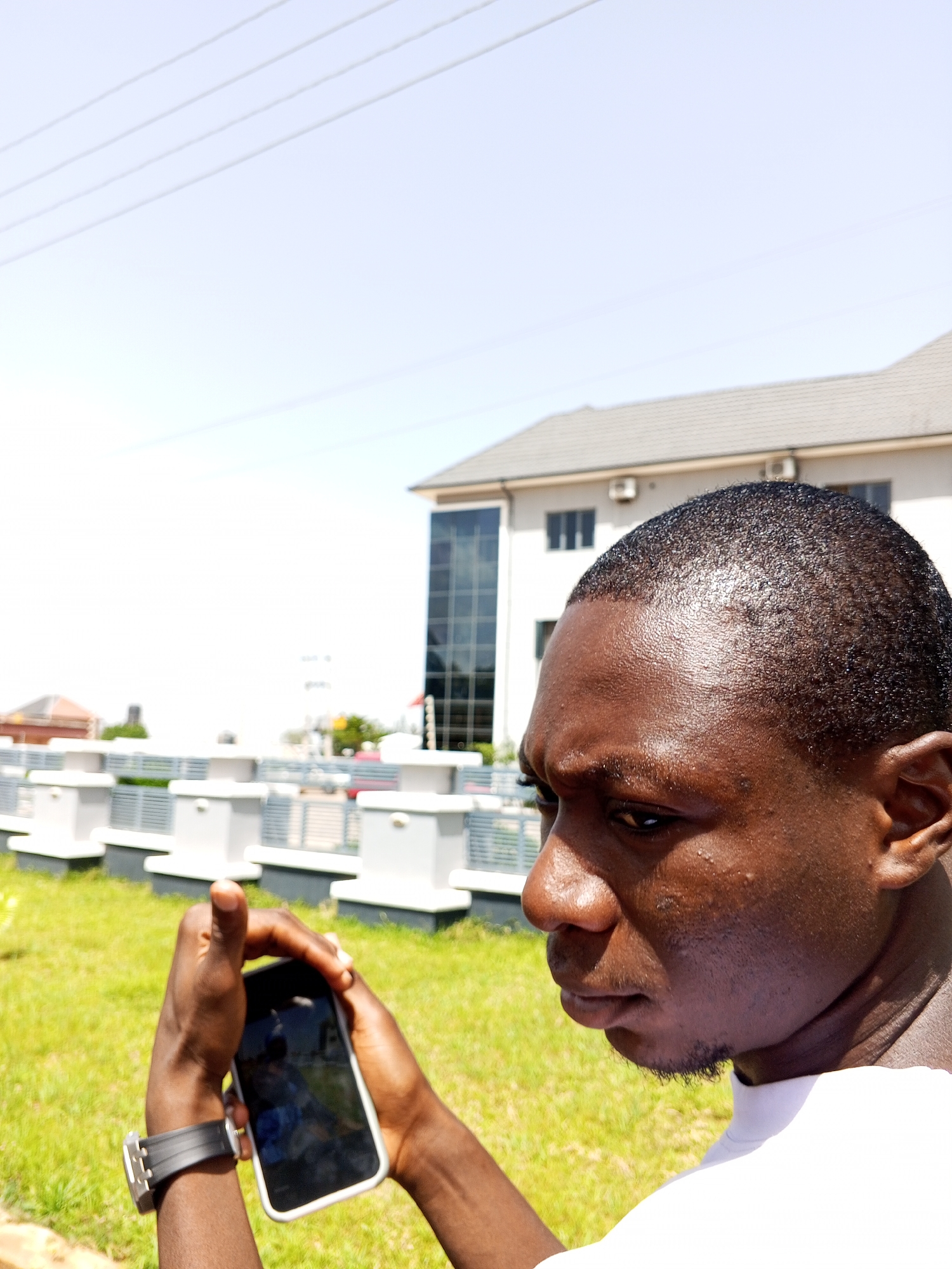 In Bukuru, Nigeria, under a bright sky, a young Black man is captured in a close-up profile shot, holding a smartphone in his hands. He is wearing a white t-shirt and a black wristwatch on his left wrist. The background reveals a modern building with glass windows and a white facade, along with a neatly manicured lawn and a series of white square structures topped with railings, suggesting an outdoor public or commercial space. The man's gaze is directed towards the phone, indicating he is engrossed in its use, possibly taking a photo or recording a video, as implied by the orientation of the device. The image focuses on the man's face and hands, with the surrounding environment slightly blurred, emphasizing the subject.
