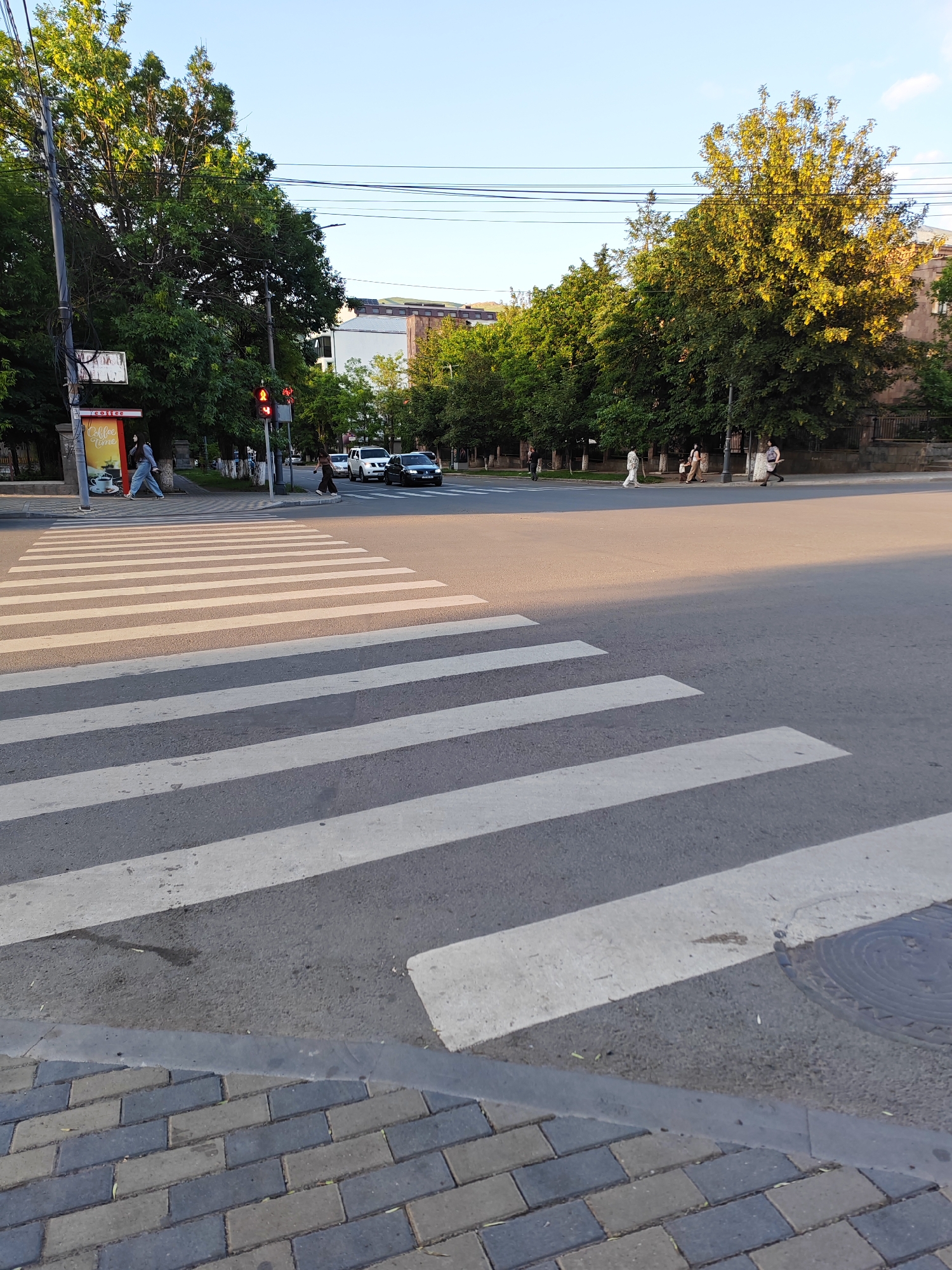 A street scene in Vanadzor, Armenia, features a wide pedestrian crosswalk dominating the foreground. The crosswalk stripes are white and clearly marked on the asphalt road. To the left, a sidewalk is paved with grey and dark grey bricks, and a short concrete curb separates it from the road.
In the background, several people are walking. Some are crossing the street on the far side of the crosswalk, while others are on the sidewalk. A traffic light is visible, currently showing red. Trees with green and yellow foliage line the street, casting shadows on the road and sidewalk. The sun appears to be low in the sky, suggesting either early morning or late afternoon, with a clear blue sky overhead.
A few cars are on the road. One black car is turning, and a white SUV is stopped. A billboard or sign is partially visible on the left, with text in what appears to be Armenian script and English. The English text includes "Coffee Time".
The overall scene depicts a typical urban street in a city, with pedestrian and vehicular traffic present. The lighting suggests pleasant weather. The presence of trees and buildings indicates an inhabited area.