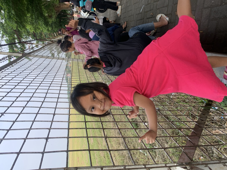 A young East Asian girl, approximately 4-6 years old, wearing a bright pink short-sleeved polo dress, stands in the foreground, facing the camera with a slight smile. She grips a weathered, rusty metal mesh fence with both hands, her thumbs visible on the horizontal wires. Behind her to the left, a flat, yellow-green grassy field extends under an overcast, light gray sky. In the far distance on the horizon, a tall, slender structure resembling an antenna or tower is visible, along with faint red-roofed buildings. To the right of the girl and behind the fence, a group of approximately 5-7 individuals, including children and adults, are gathered along a paved path, also looking towards the field. One person in a dark hooded jacket sits on the ground, wearing blue jeans and white flip-flops, with their left arm raised in a thumbs-up gesture. Another child in a light pink top holds the fence. An adult male in a brown cap and purple shirt is partially visible behind them. Two motorcycles, one yellow and one dark, are parked behind the group. Green trees line the paved path in the background. The scene is set outdoors in the city of Desa Ngesrep, Indonesia.