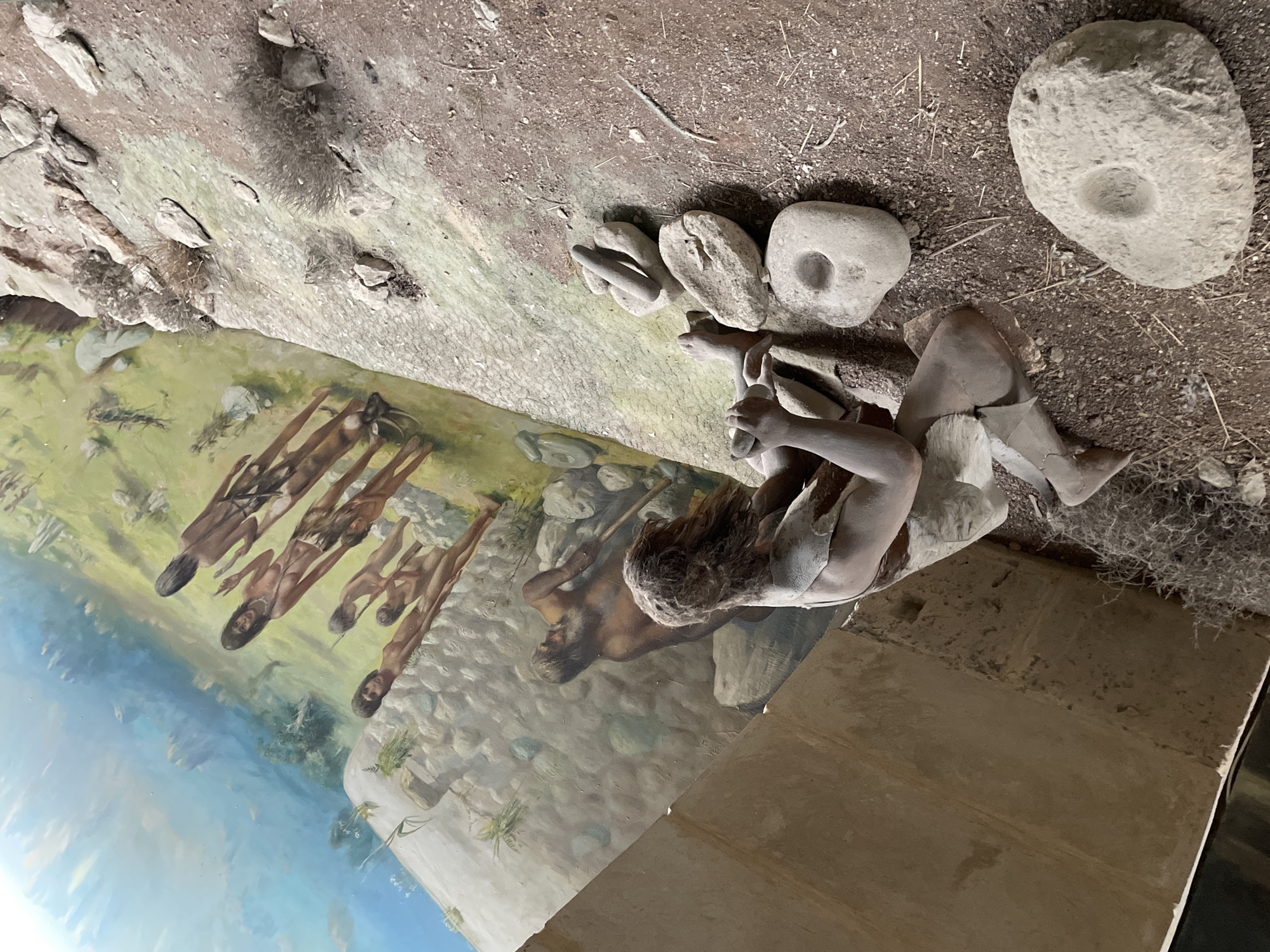 The image depicts a diorama representing a prehistoric scene, likely a museum exhibit. In the foreground, a full-body mannequin of a person is kneeling, engaged in an activity that involves grinding stones. The mannequin is dressed in a loincloth and has a primitive aesthetic. Surrounding the mannequin are several stone tools, including grinding stones with depressions and a pestle-like object. The background features a painted mural depicting a group of people, possibly hunters or gatherers, in a landscape. This mural is part of a larger environmental display, with rock formations and sparse vegetation visible. The overall scene is presented in a slanted perspective, giving the impression of looking down onto the exhibit. The location is identified as the Kyrenia Castle Museum in Kyrenia, Northern Cyprus.