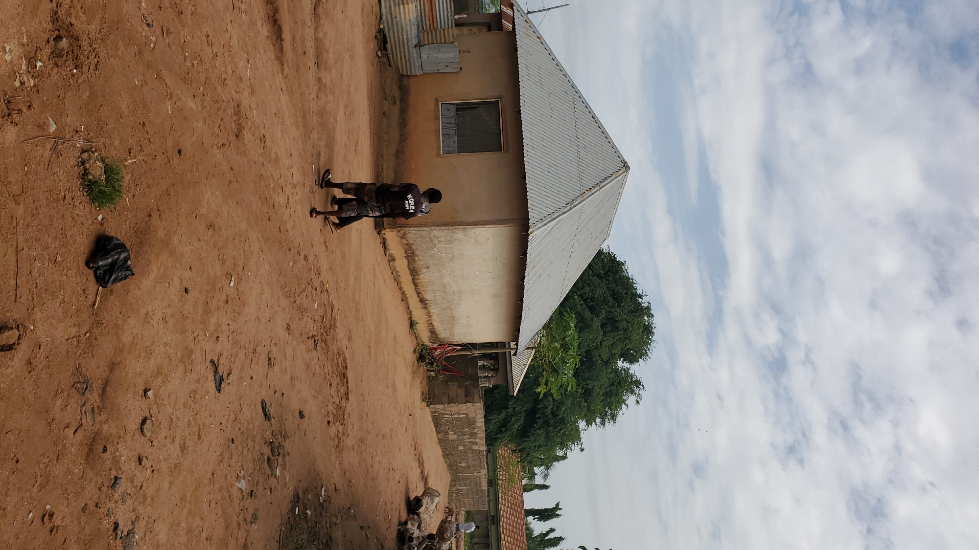 The image depicts a dusty, unpaved street in a residential area, likely in the city of Jalingo, Nigeria, under a cloudy sky. In the foreground, two young males are walking away from the camera. The one in front, on the left, wears a black t-shirt with "KOREA ARMY 2017" printed on the back and dark shorts. The second male, slightly behind and to the right, is mostly obscured but appears to be wearing dark shorts. A black plastic bag of trash lies on the ground near the center of the frame.
To the left of the street, a light-colored, rectangular house with a corrugated metal roof is visible. It has a single window with a patterned covering. To the left of this house, there are sections of corrugated metal sheeting, possibly part of another structure or fencing.
Further down the street, a low, rough-hewn stone wall runs along the right side. Beyond the wall, more houses with tiled roofs and some lush green trees can be seen. One of these trees, a large, dense canopy, is prominent behind the first house. In the distance, several tall, thin trees with distinctive crowns dot the landscape. The ground is predominantly reddish-brown earth, with sparse patches of dry grass and scattered debris. The overall impression is of a simple, rural or semi-urban environment.