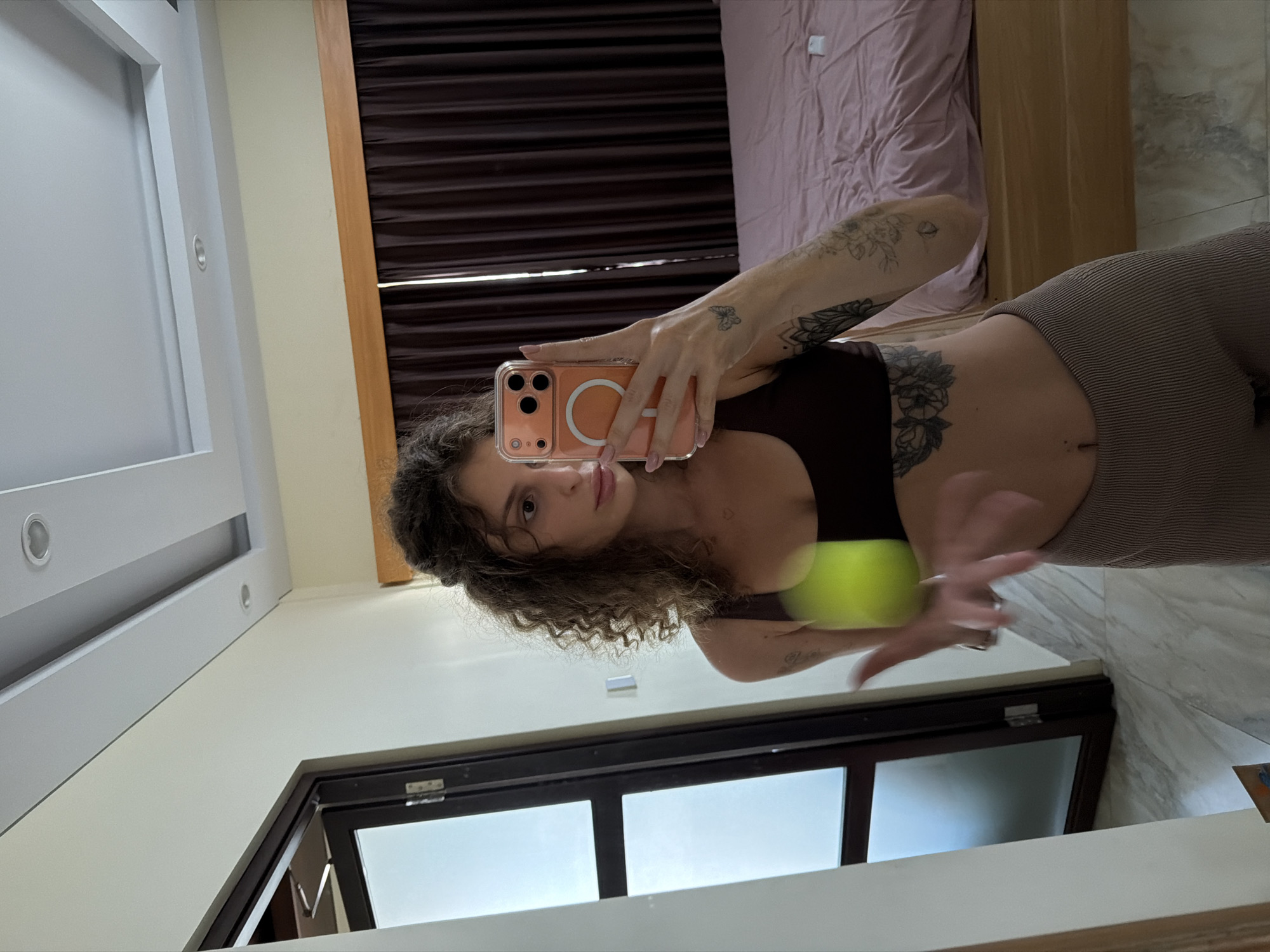 The image captures a young woman taking a mirror selfie inside what appears to be a bedroom or hotel room, potentially in North Kuta, Indonesia. The woman has curly dark brown hair and is wearing a dark-colored sports bra or crop top and light brown/khaki ribbed leggings. She holds a peach-colored iPhone with a clear case and a visible white MagSafe ring, which partially covers her face as she looks directly into the camera. Her hands have light pink nail polish, and she displays multiple tattoos, including a floral design on her lower torso/hip, a butterfly on her right hand, and other patterns on her left arm. A blurry, neon green object, possibly a ball, is held in her left hand.
The room's setting includes white walls, a white ceiling with recessed lights, and a light-colored tiled or marble floor. In the background, a bed with light pink bedding and a wooden frame is visible. Dark brown horizontal blinds or slatted wall panels are behind the bed. A window with dark frames is present towards the bottom of the image. The overall impression is a modern and clean indoor environment during the daytime. No visible text is discernible in the image.