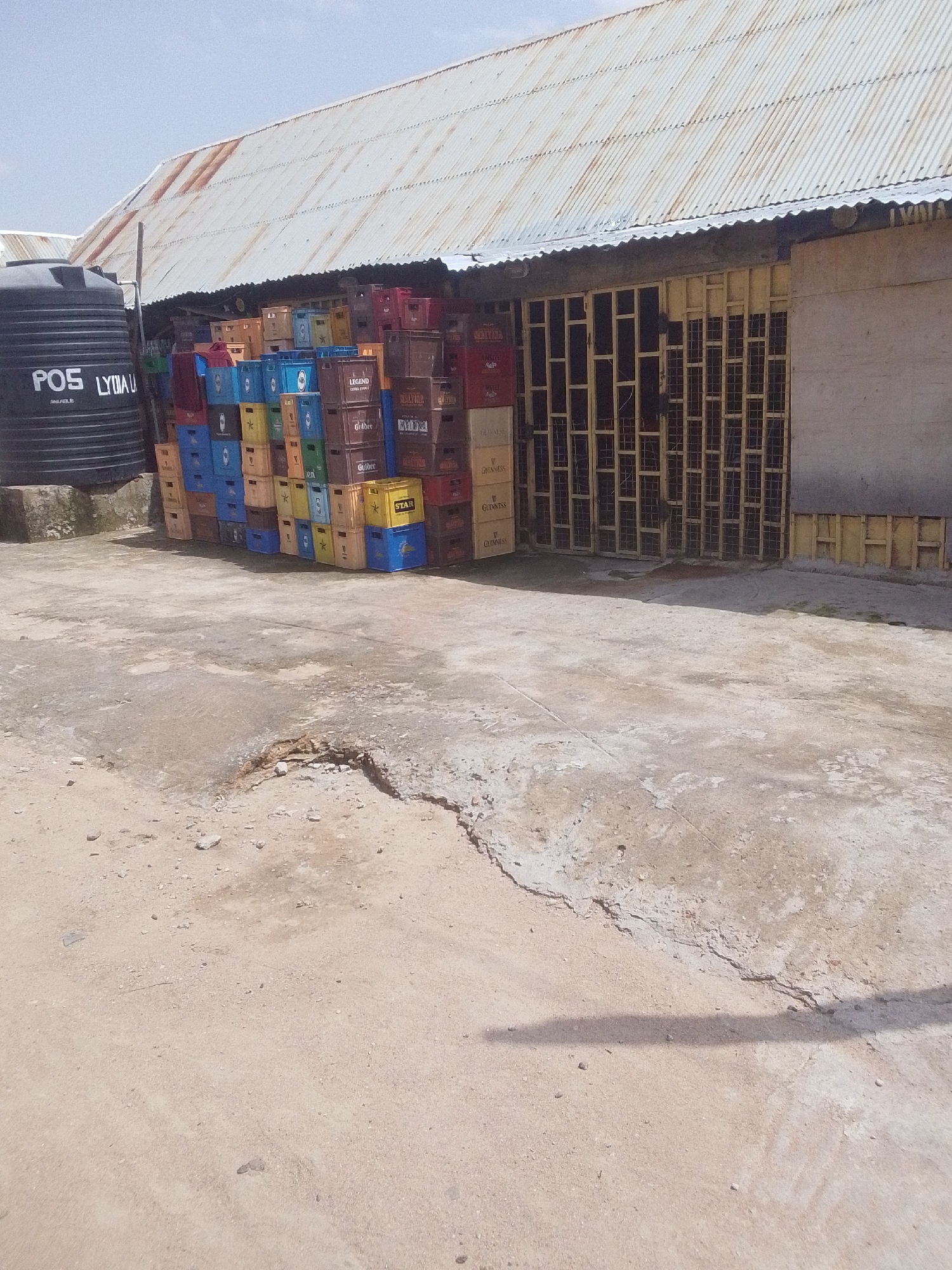 The image shows a pile of empty beverage crates stacked outside a building with a corrugated metal roof. A large black water tank with "POS LYDAU ANLABLE" printed on it is visible to the left of the crates. The building appears to be a shop or storage area, with a barred window and door. The ground is concrete and dirt, with some cracks and debris. The crates are various colors, including red, blue, yellow, and brown, and some have labels such as "LEGEND," "Gulder," "Guinness," and "STAR." The scene suggests a commercial or retail setting, possibly a distribution point for beverages. The lighting and shadows indicate it is daytime, and the sky is clear, suggesting fair weather. The location context of Jalingo, Nigeria, implies a warm climate. There are no people visible in the image, so no activity is directly observed, but the presence of the crates suggests a business that deals with beverages. The overall impression is one of a functional, perhaps slightly unkempt, commercial space.