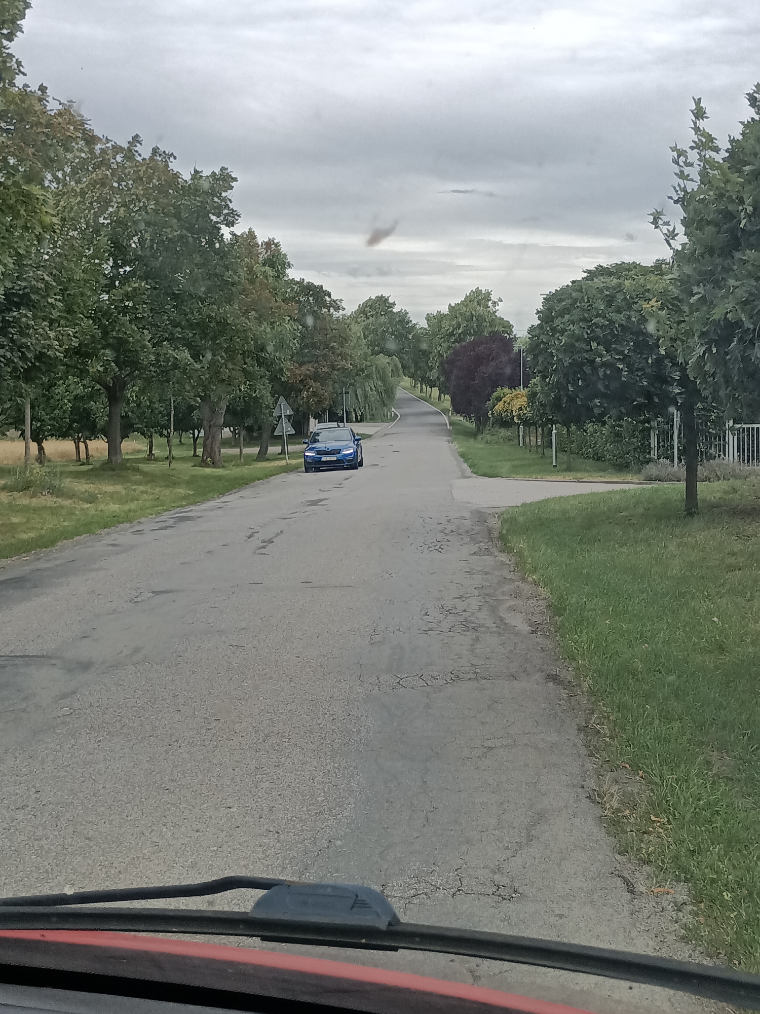 This is a photograph taken from inside a vehicle, looking out at a rural road in Svémyslice, Czechia. The scene depicts a slightly overcast day, likely in the late morning or afternoon, given the diffused natural light. The road is paved but shows signs of wear with cracks and patches.
The main subject in the scene is a bright blue car driving away from the camera on the right side of the road. Trees line both sides of the road, with lush green foliage indicating a season like summer or early autumn. On the right, there's a white fence and manicured grass, suggesting a residential or cultivated area. On the left, the landscape is more natural, with a field bordering the trees.
The activity is simply driving on a road. There are no people visible in the frame. Notable details include the overcast sky, the condition of the road, and the variety of trees, some of which are larger and more mature, while others appear to be more ornamental, like the clipped trees along the right side. The perspective from within the vehicle is evident from the dashboard and windshield wipers visible at the bottom of the frame. The overall mood is calm and mundane, depicting a typical drive through the countryside. No visible text is present in the image.