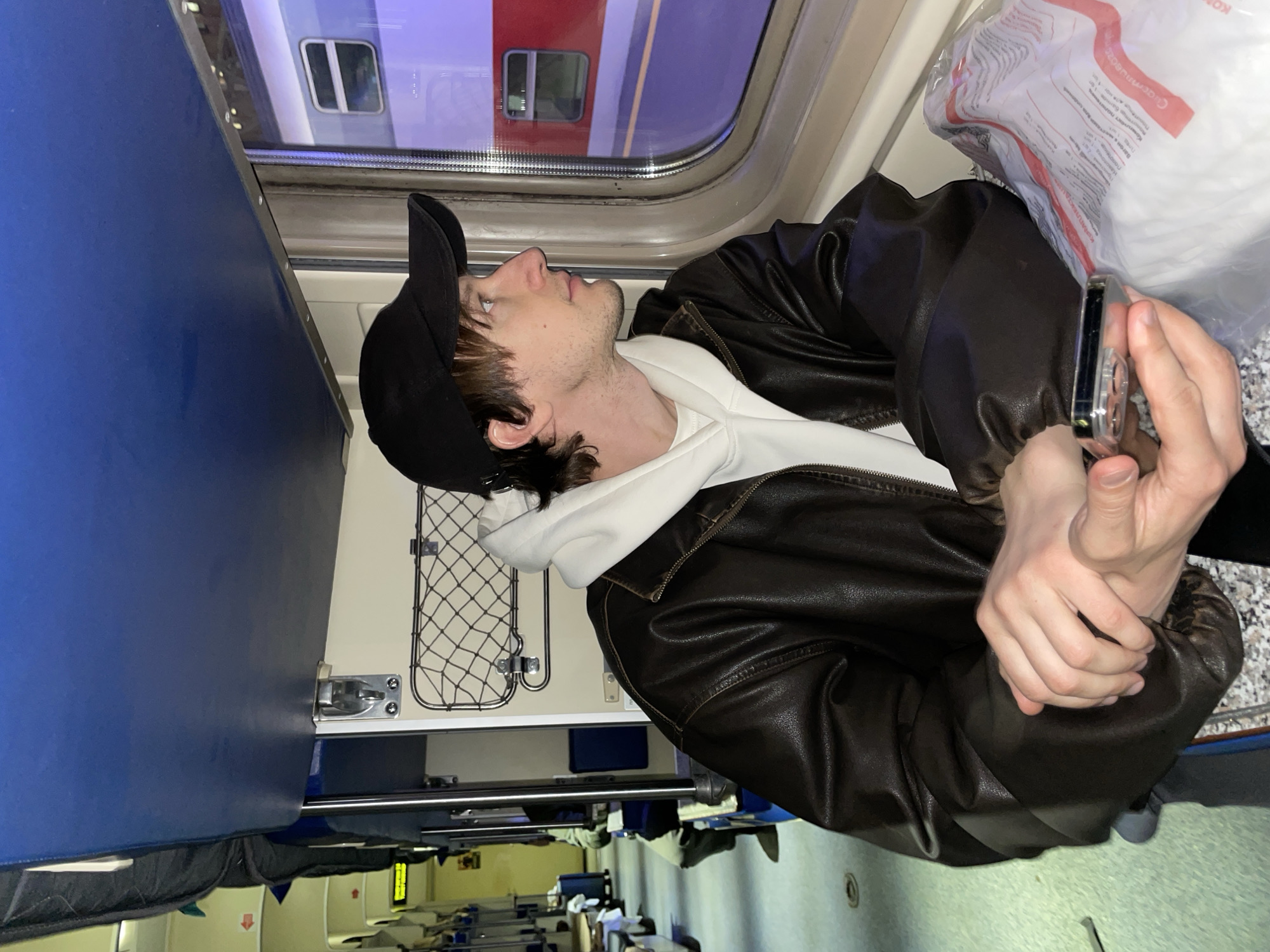 A person wearing a black baseball cap, white hoodie, and brown leather jacket is looking up at the window of a train. The person is holding a smartphone in their hands. Outside the window, a red train is visible. The interior of the train compartment includes blue seating and a luggage net. The city and location are not discernible from the image.