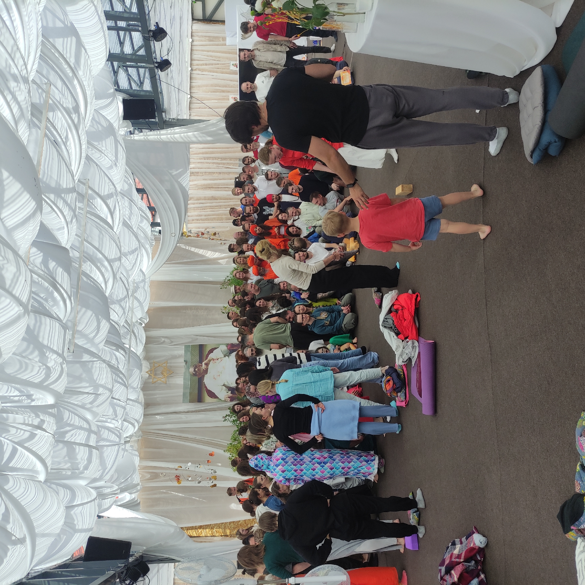 The image depicts a gathering of approximately 50 people in an indoor or semi-indoor space covered by white draped fabric. The attendees are of various ages and genders, dressed in casual attire. Some are seated on the floor, while others are standing. In the background, a large portrait of a man in white is displayed. The environment appears to be set up for an event, possibly a workshop or gathering, with yoga mats and blankets visible on the floor. The lighting is diffused, suggesting natural light supplemented by artificial sources. The overall atmosphere seems relaxed and communal. This scene is taking place in Zhirnosovo, Russia.