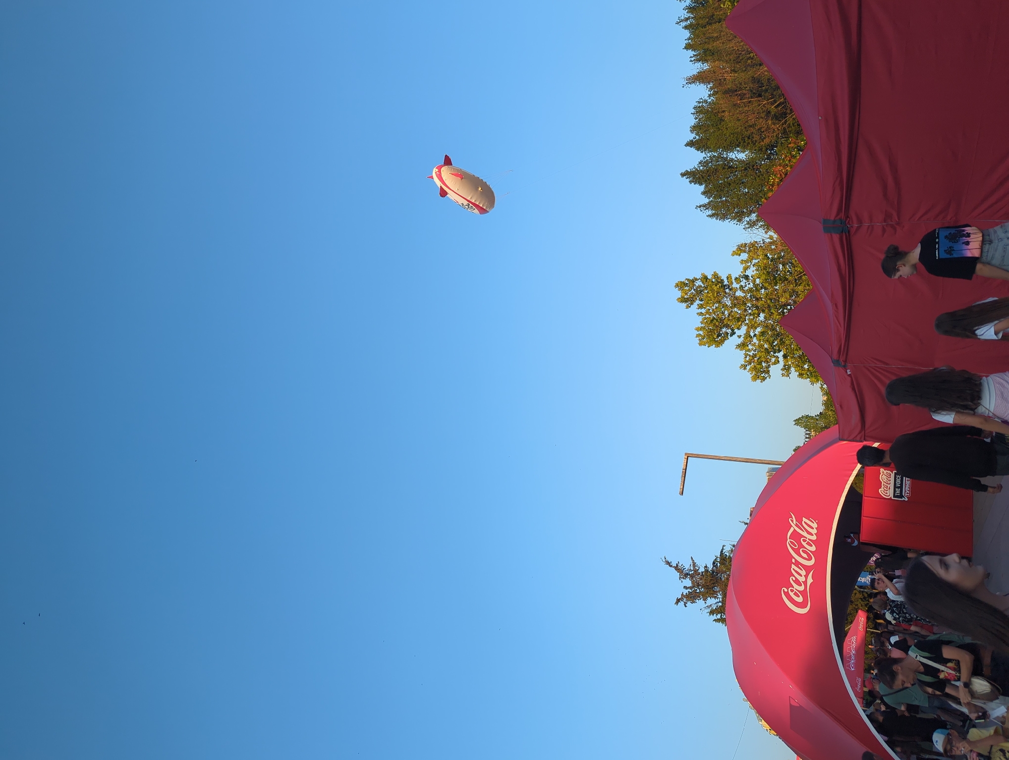 An outdoor event is in progress under a clear blue sky. In the foreground, a large, arched, red Coca-Cola branded tent is visible, with people entering and exiting. Behind this tent, a row of smaller, red marquees stretches across the scene. A blimp, also branded with Coca-Cola and featuring red accents, floats high in the sky. Trees are visible in the background, and several small birds are scattered across the sky. The event appears to be a public gathering.