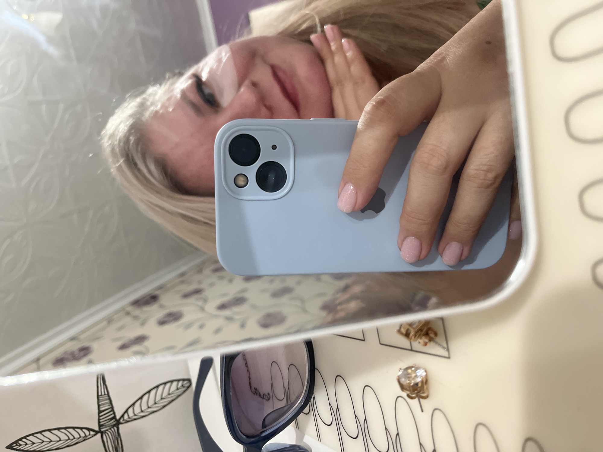 The image is a selfie taken in a mirror. A woman with blonde hair is partially visible in the reflection. She is holding a light blue iPhone in her right hand, which is covering part of her face. The phone has three camera lenses. The mirror frame is white. Below the mirror's reflection, there is a pair of sunglasses with dark frames, and two gold-colored earrings on a white surface with black oval-shaped designs. The background is out of focus, but a floral patterned surface is visible. There is no evidence to support the claim that the image was taken in Izhevsk, Russia.