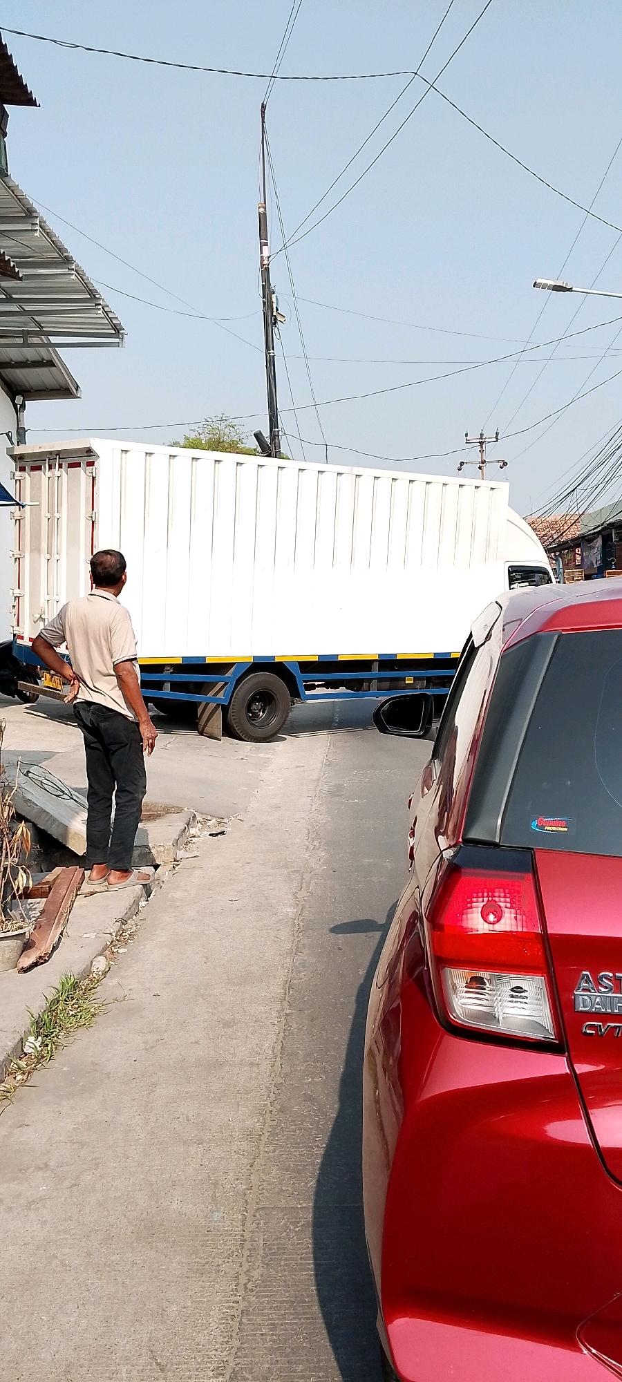 The image captures a daytime street scene in Babelan, Indonesia. A large white container truck is parked or moving slowly on the right side of the road. To the left of the truck, a man in a light-colored short-sleeved shirt and dark pants stands facing away from the camera, looking towards the truck. He appears to be standing on a concrete sidewalk, which is uneven and shows some signs of wear. A red car is visible in the foreground on the right, partially obscuring the view of the truck and road. The environment includes overhead power lines and a building with a metal awning on the far left. The sky is clear and blue.