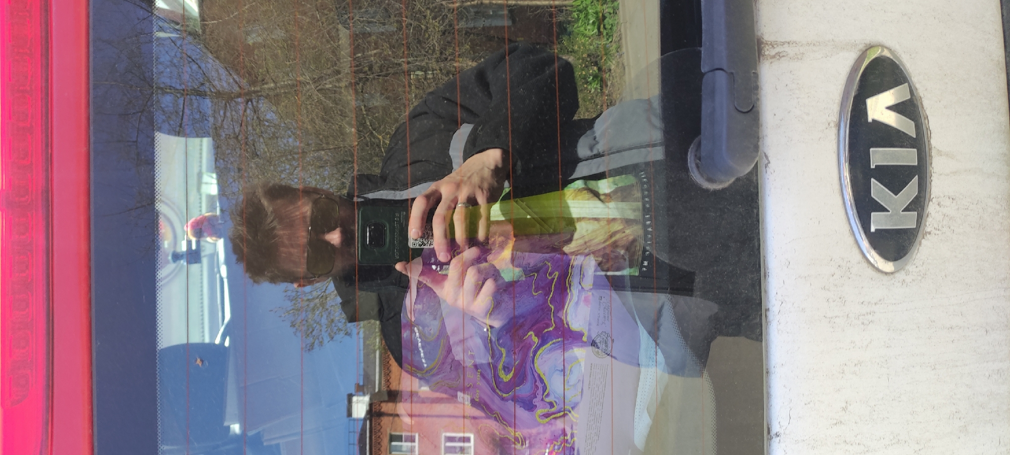 A person is taking a photo of the back of a Kia car. The car is light-colored and has a rear windshield wiper. The person is reflected in the car's rear window, and they are wearing a dark jacket and sunglasses. They are holding a smartphone to take the photo. There is a reflection of a building in the rear window, which places the location in Fryazino, Russia.
