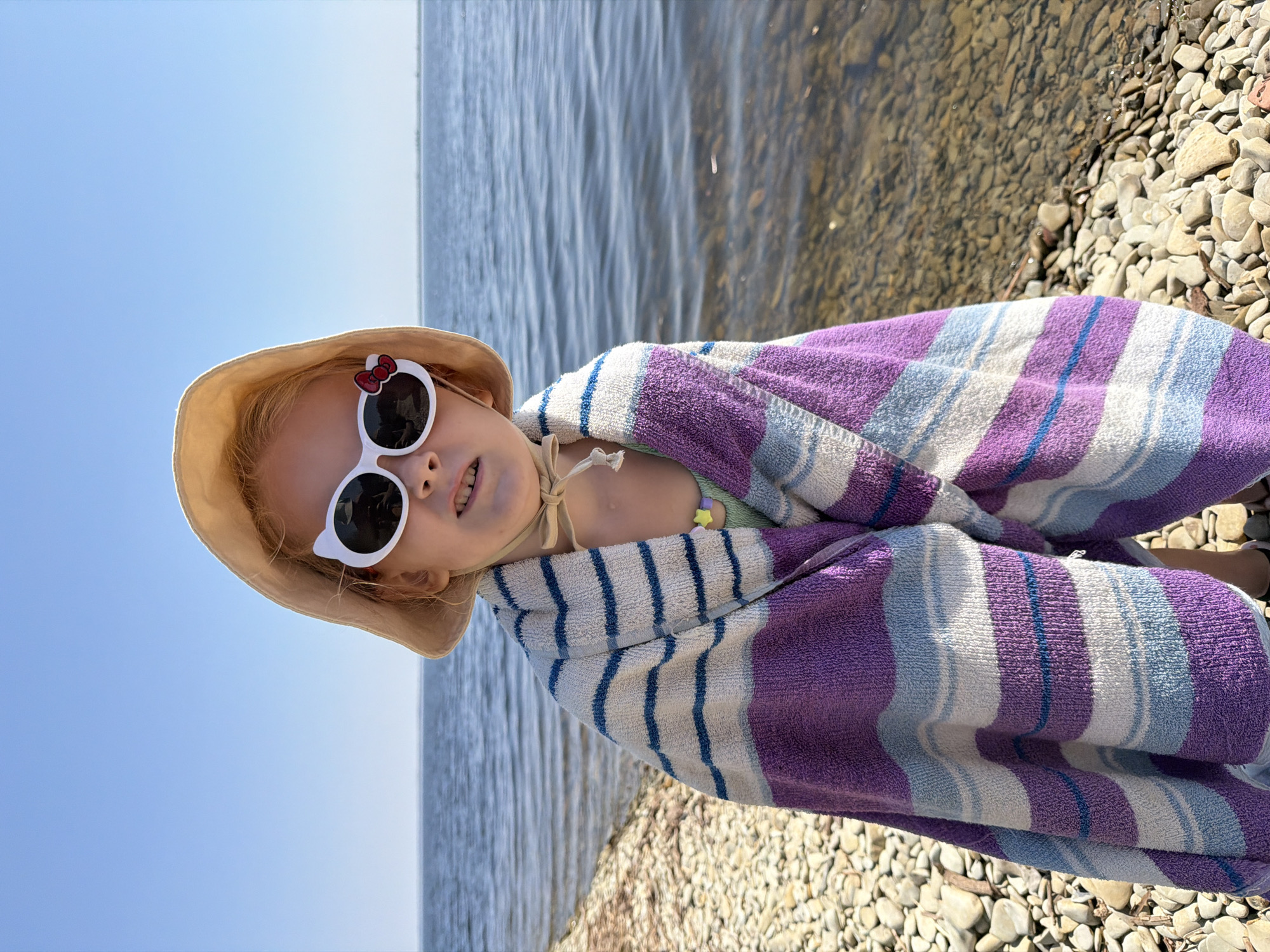 A young girl is wrapped in a striped towel on a rocky beach. She is wearing a sun hat and white sunglasses with red bows. The water is calm and appears to be a lake or river. The sky is clear and blue, suggesting a sunny day. The pebbles on the beach are smooth and varied in color. The girl is looking at the camera with a slight smile, and her body language suggests she is enjoying the outdoors. The scene evokes a sense of summer vacation and relaxation. It is daytime, and the weather is warm and pleasant. The location is a beach, likely near a body of water. The girl's attire and the beach setting suggest a recreational activity like swimming or playing near the water. There is no visible text in the image.