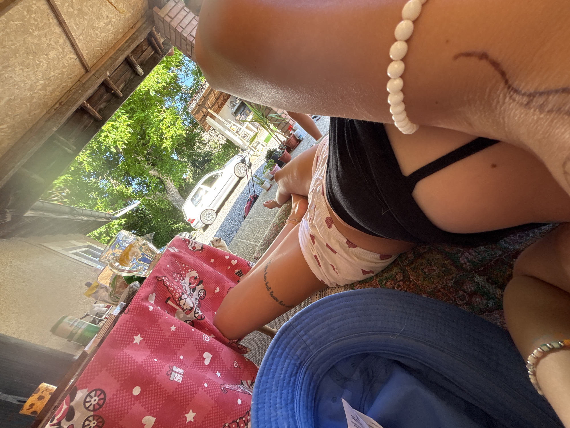 The image captures a sunlit outdoor scene, likely a backyard or patio area, with a person lying down. A blue bucket hat is placed on the left side of the frame. In the center, a person's legs are visible, wearing patterned shorts and a dark bikini bottom. A tattoo is visible on their left thigh. To the right, another person's arm and shoulder are in the foreground, adorned with a white beaded bracelet. A table with a pink tablecloth featuring white stars and cartoon gnomes is in the background. On the table, there are drinks and various items. A white car is parked in the driveway in the distance. The environment appears to be a residential area with trees and a partial view of a building. The location is identified as Belopoptsi, Bulgaria.